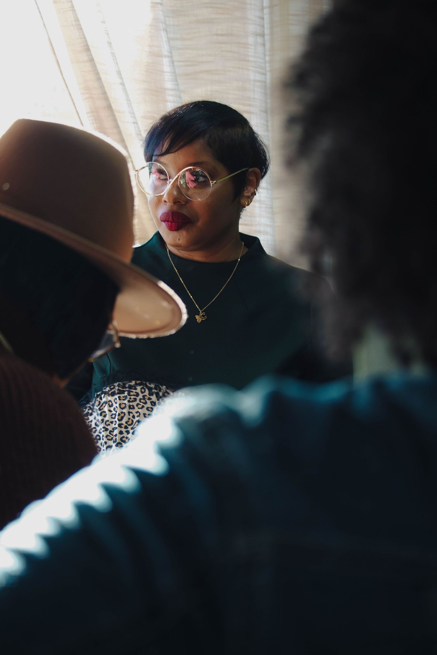 Stylish Black woman with short hair, red lips, and glasses seen between two blurred figures indoors.