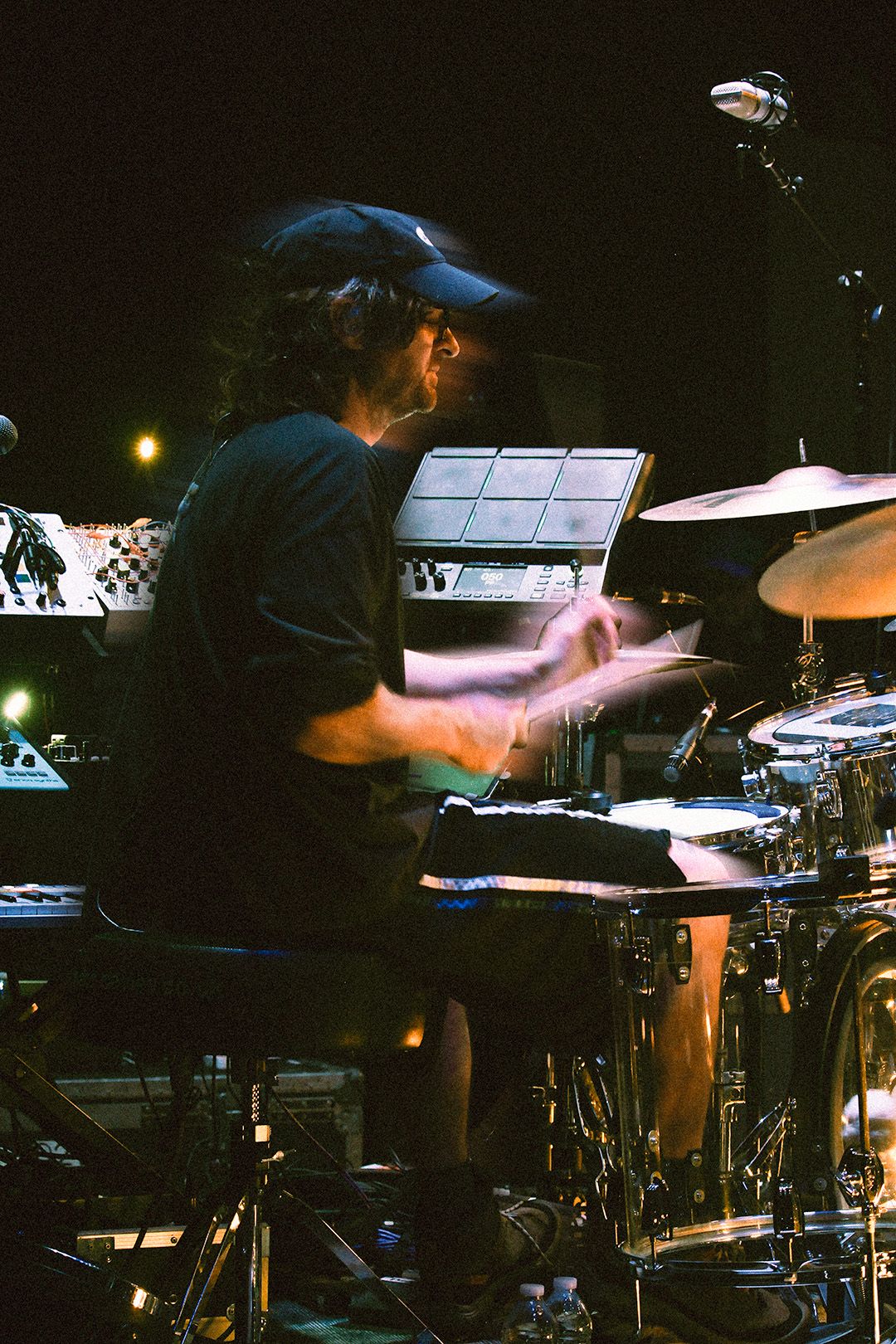 Drummer in baseball cap plays drum kit energetically on a dark stage with motion blur on hands.