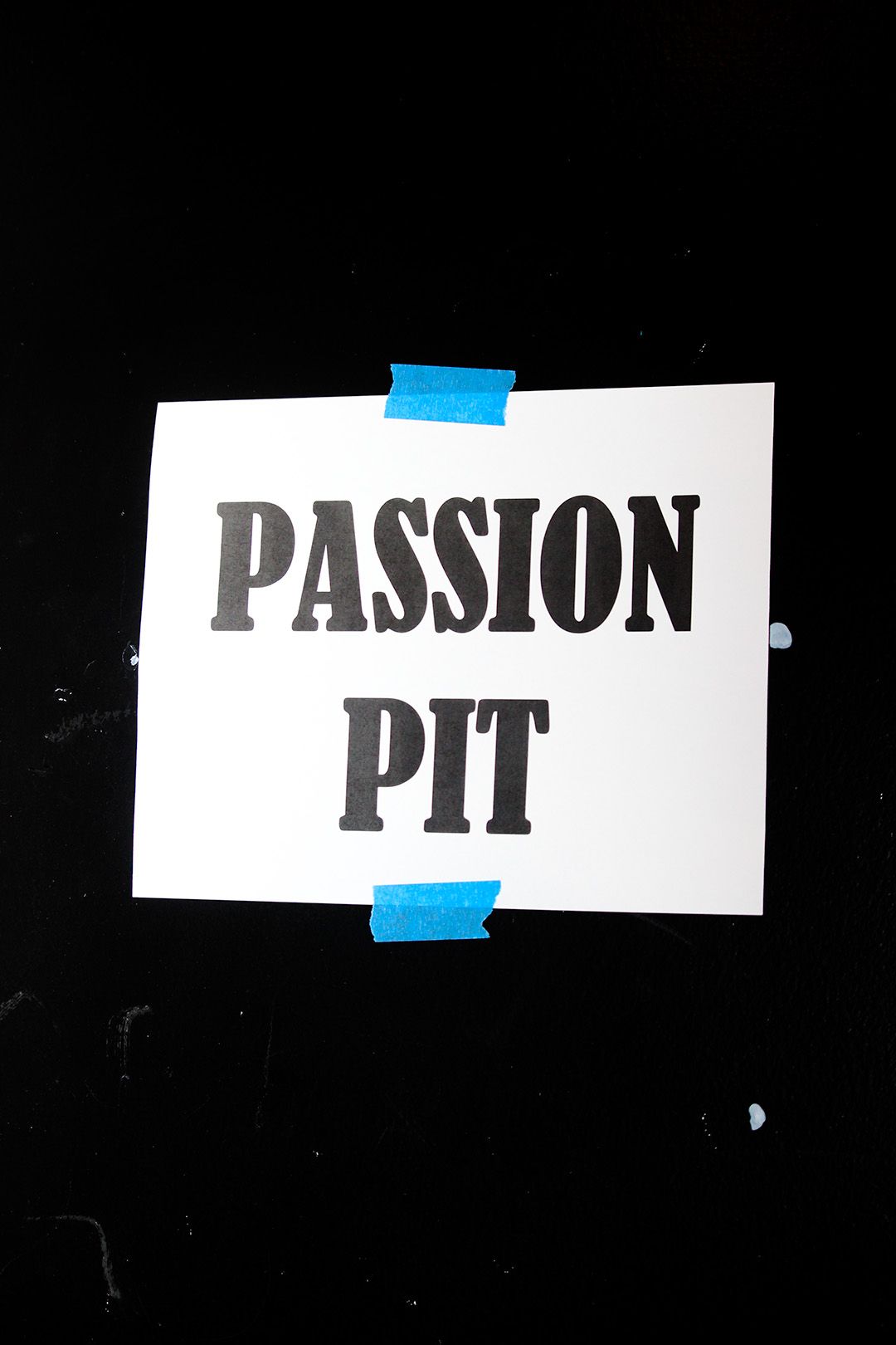 White paper sign reading Passion Pit taped with blue tape on black wall in dark venue.