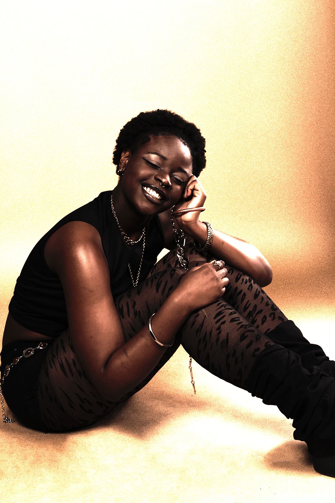 Black woman with short natural hair smiles while sitting on floor in black outfit with chain accessories.