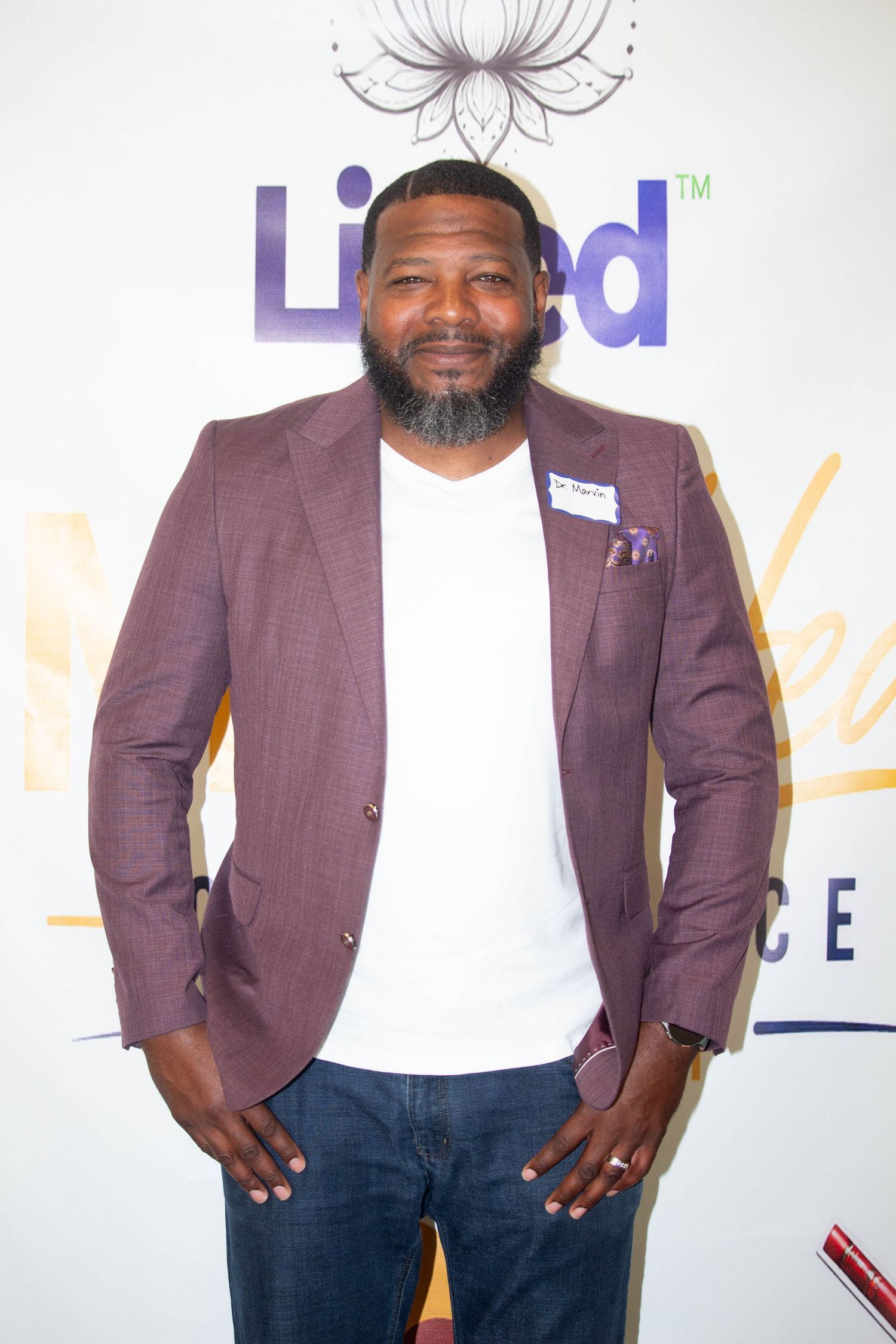 Man in purple blazer and white t-shirt posing at a branded event backdrop, smiling with hands in pockets.