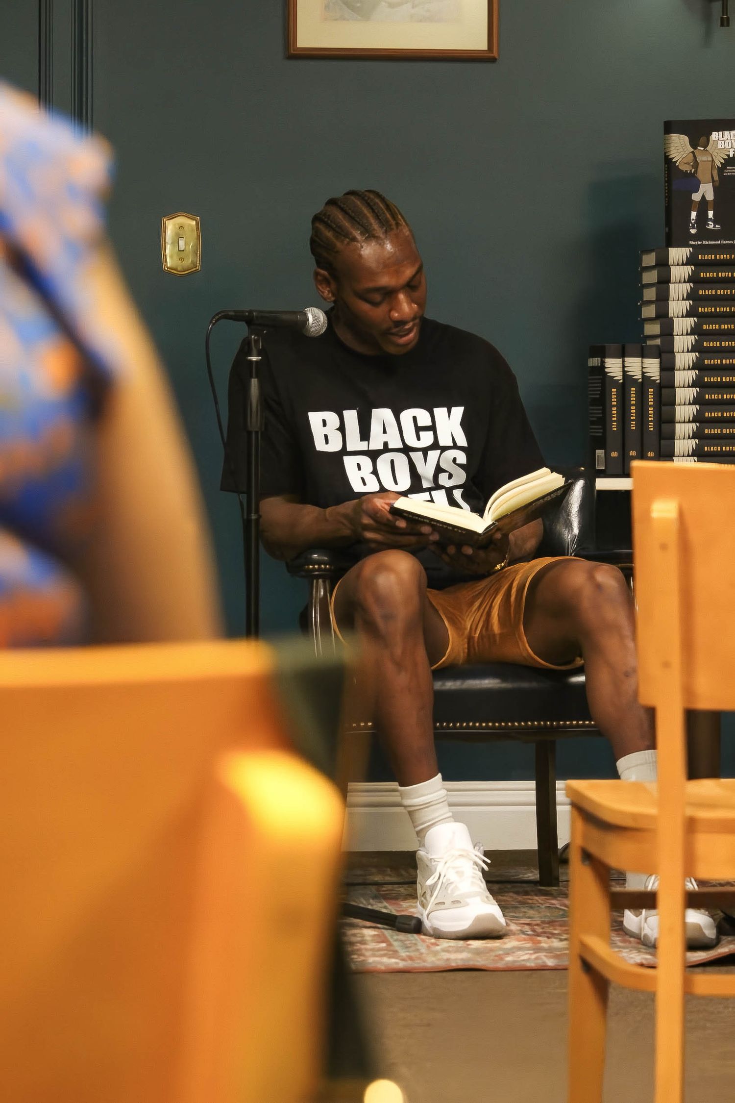 Man in Black Boys Read shirt sits reading a book near a microphone at a bookstore event.
