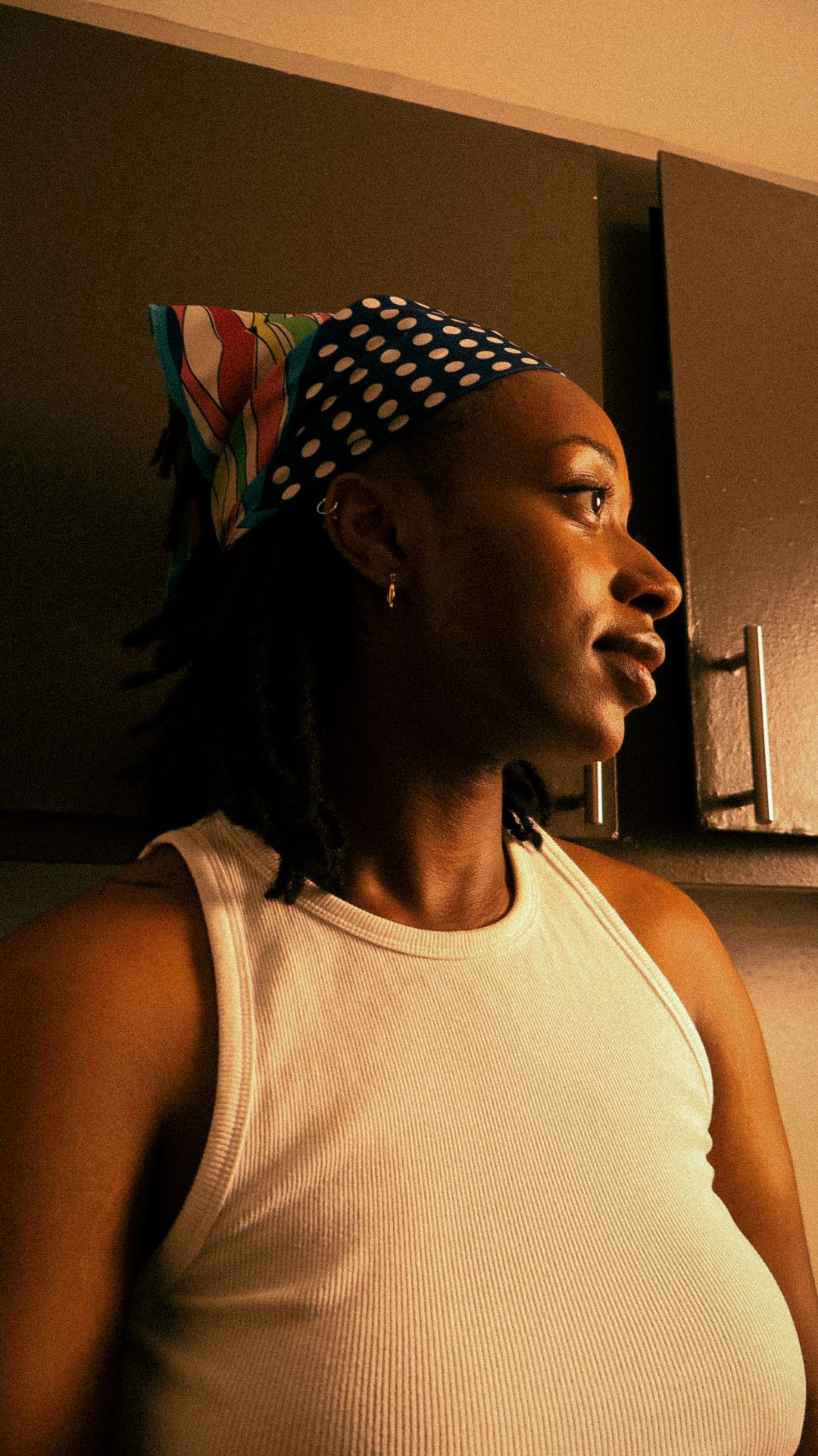 Young woman wearing a polka dot headband and white tank top standing in a dimly lit room.