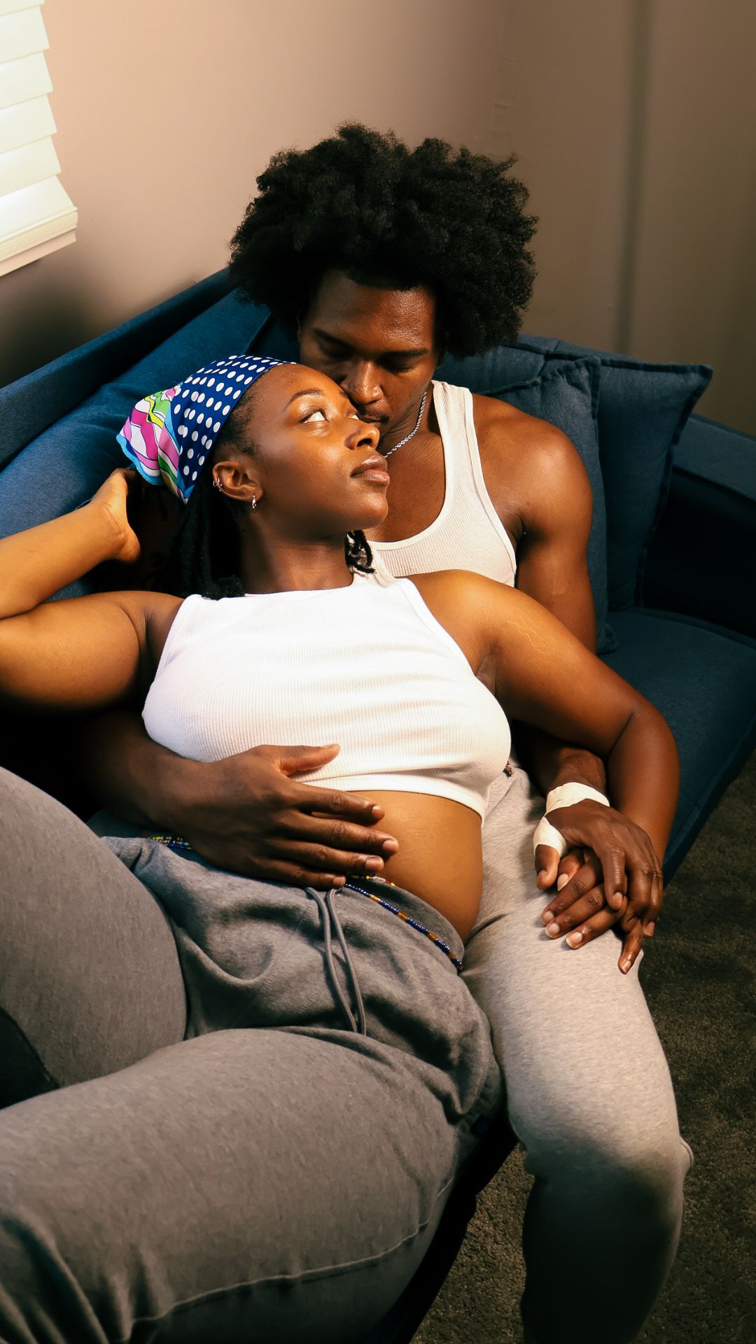 A young Black couple cuddles intimately on a blue sofa, woman leaning back against mans chest.