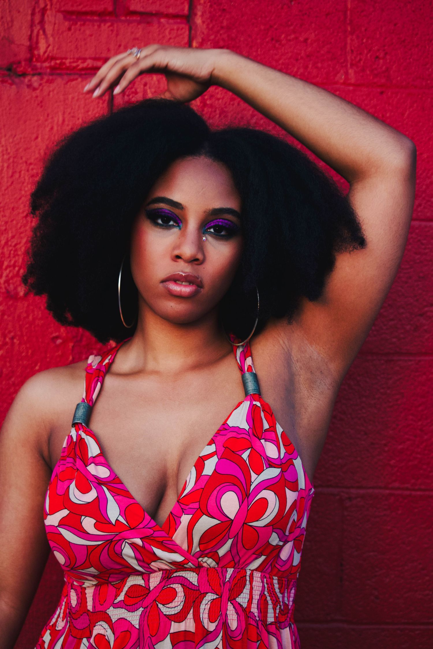 A young Black woman with an afro and purple eye makeup poses in a colorful pink floral dress against a red wall.