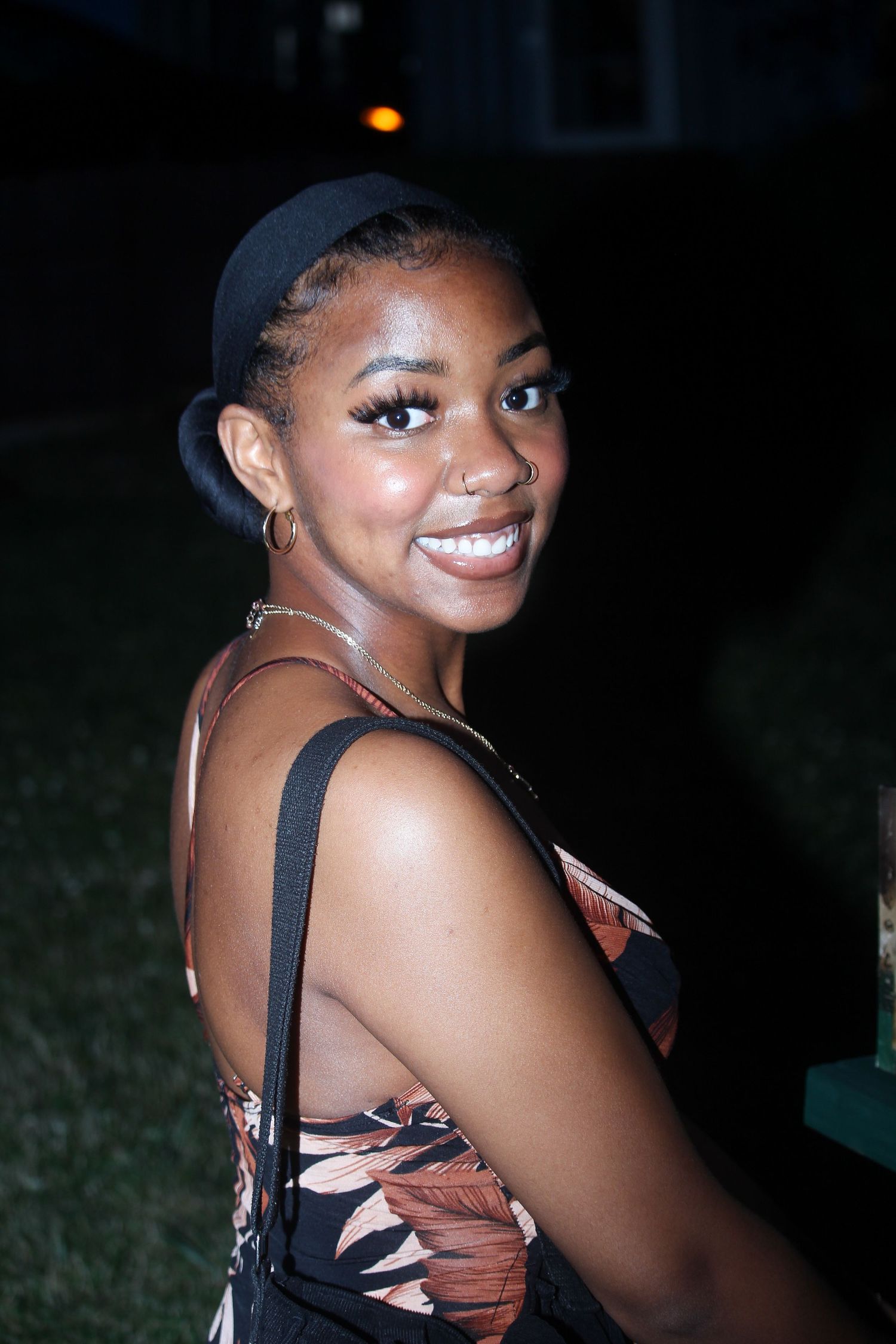 Smiling young woman in floral dress glances back over shoulder at an outdoor nighttime event.