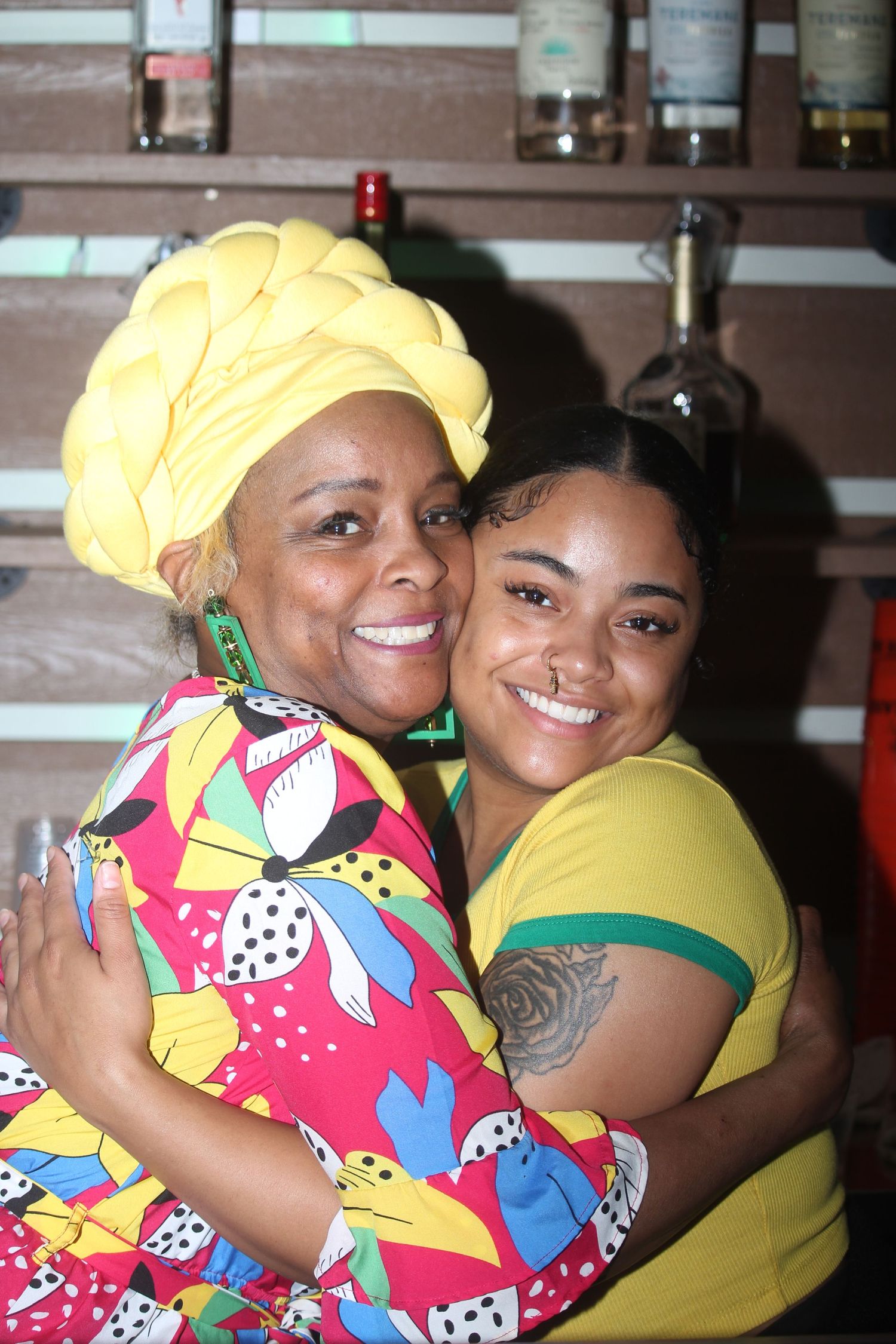 Two women share a warm hug, one wearing a vibrant yellow head wrap and colorful dress at a bar.