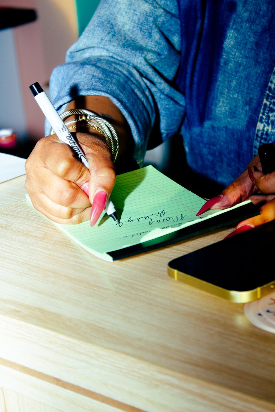 Close-up of a woman with pink nails writing on a green notepad at a wooden desk.