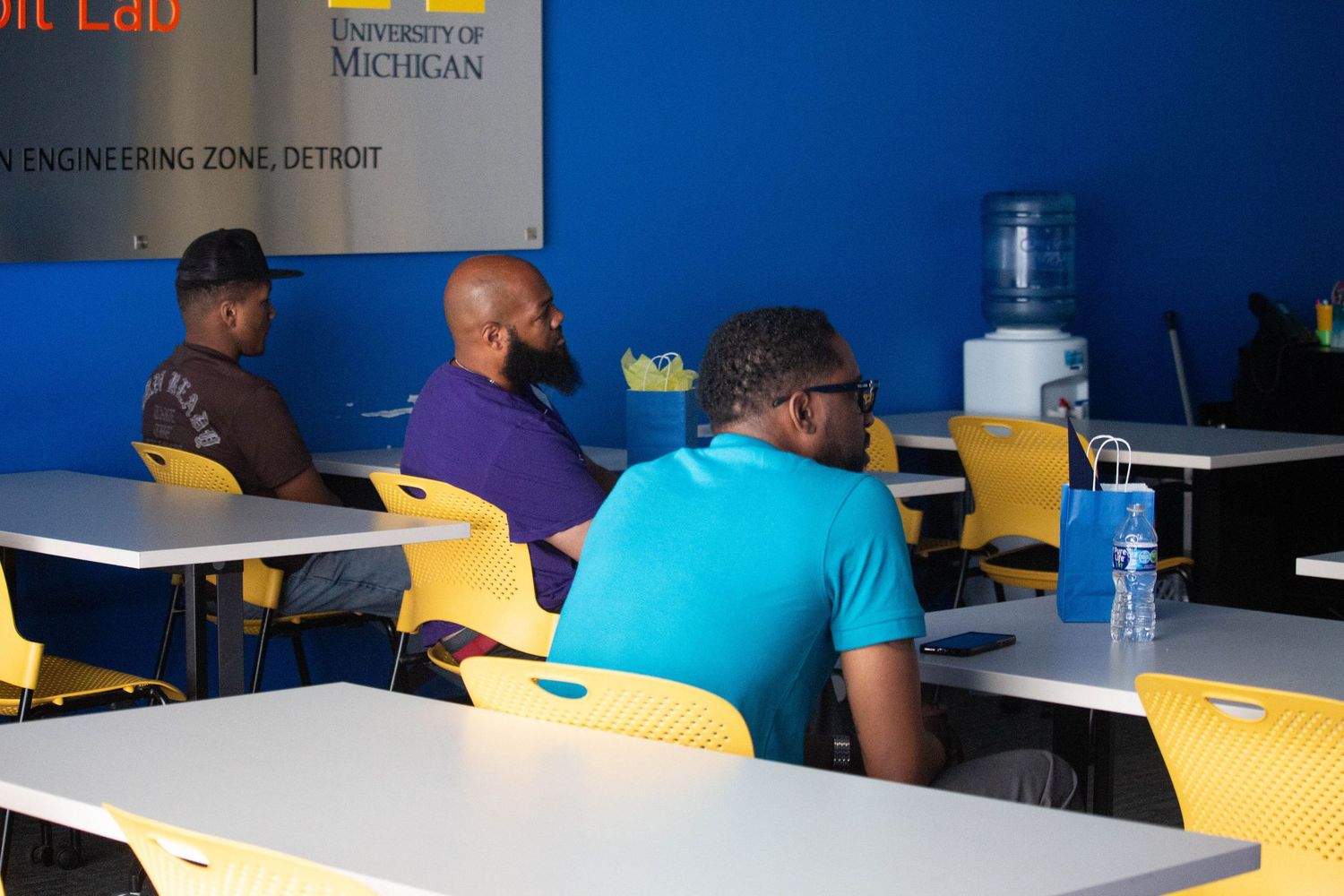 Three men sit at yellow chairs in a University of Michigan Engineering Zone classroom in Detroit with a water cooler nearby.