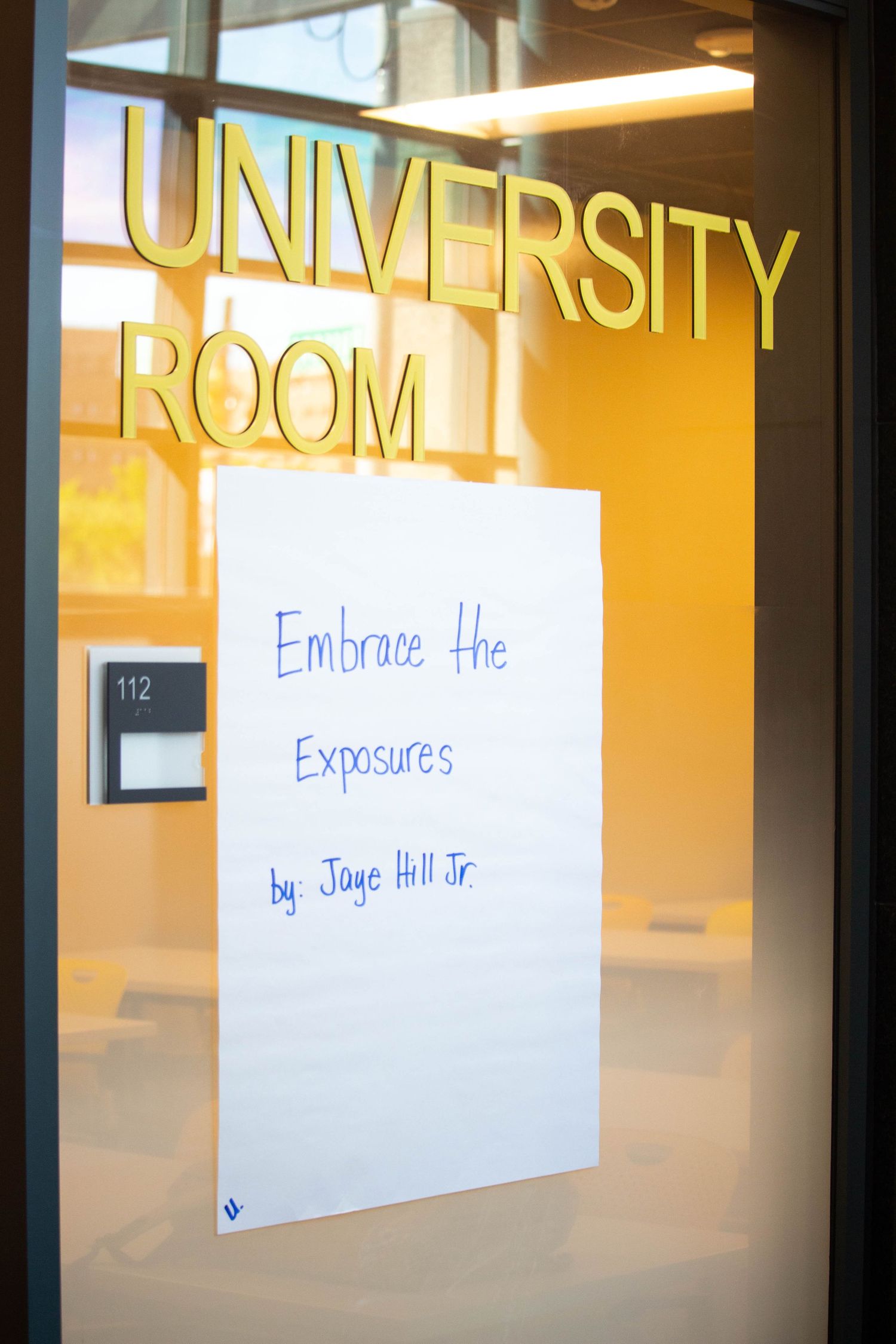 University Room door with a sign reading Embrace the Exposures by Jaye Hill Jr. at room 112.