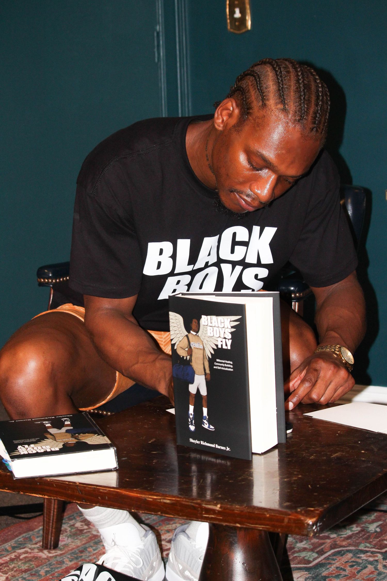 Man in Black Boys t-shirt signing copies of Black Boys Fly book at a wooden table.