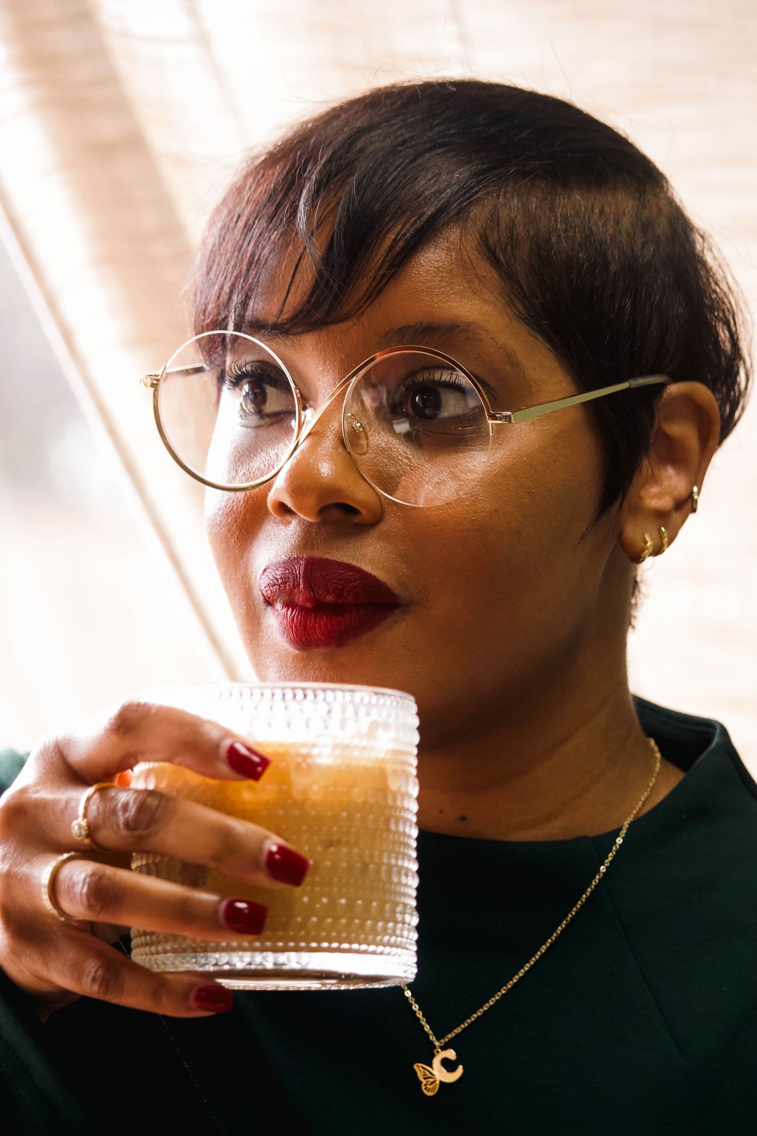 Stylish woman with short hair, round gold glasses, red lips, holding a craft cocktail at an author signing event.