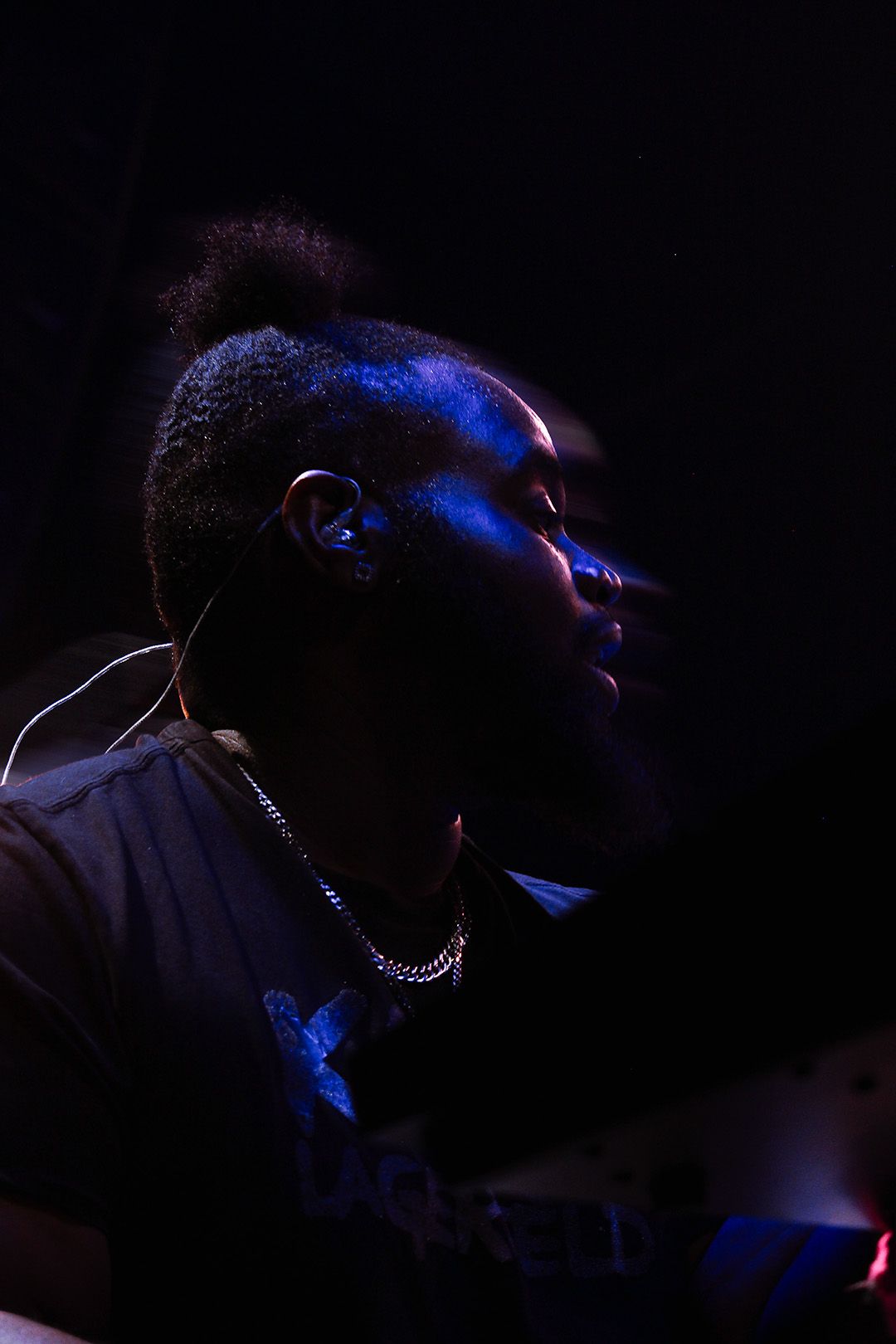 Black male performer lit by blue light, wearing earpiece and chain necklace, eyes closed on dark stage.