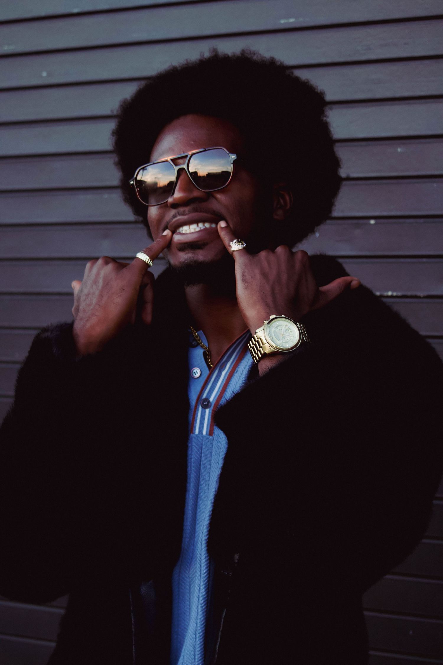 Smiling Black man with afro wearing sunglasses, gold watch, and black fur coat posing against a dark wall.