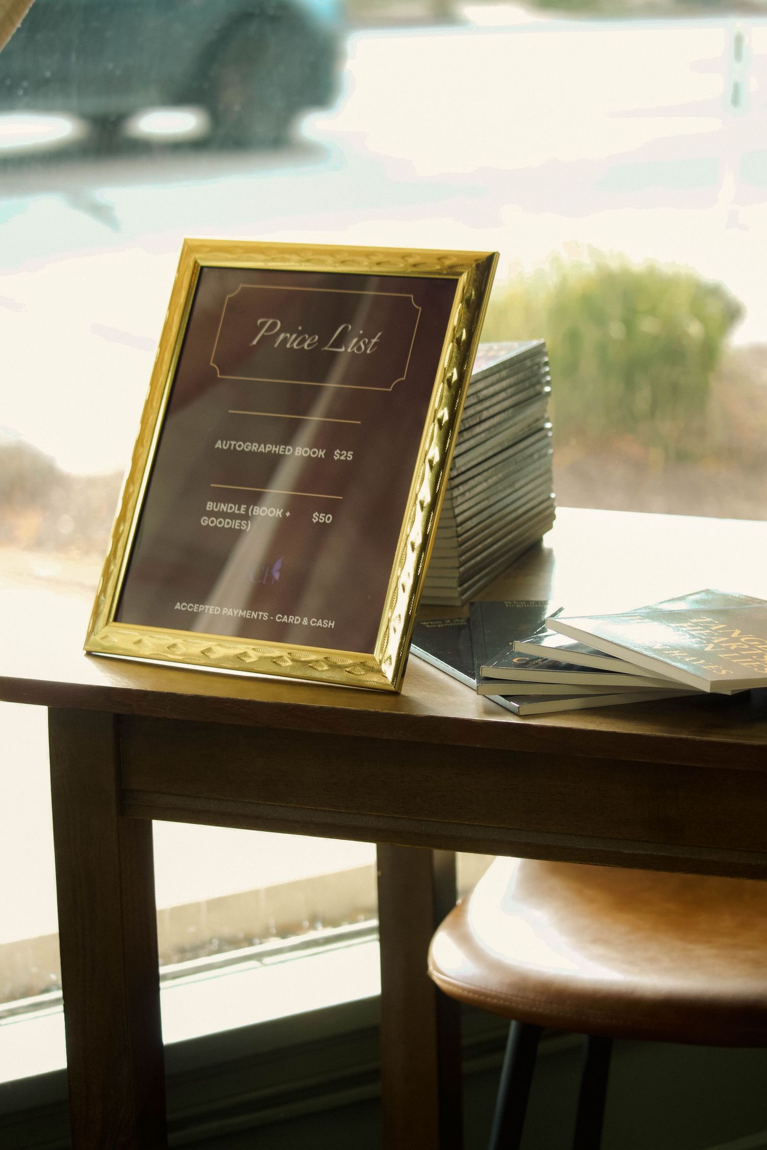 Gold-framed price list sign beside a stack of autographed books on a table at a Carmen Hayes book signing.