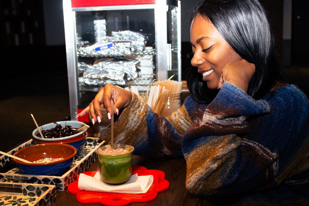 Smiling woman stirring a green matcha drink at a table with toppings nearby.