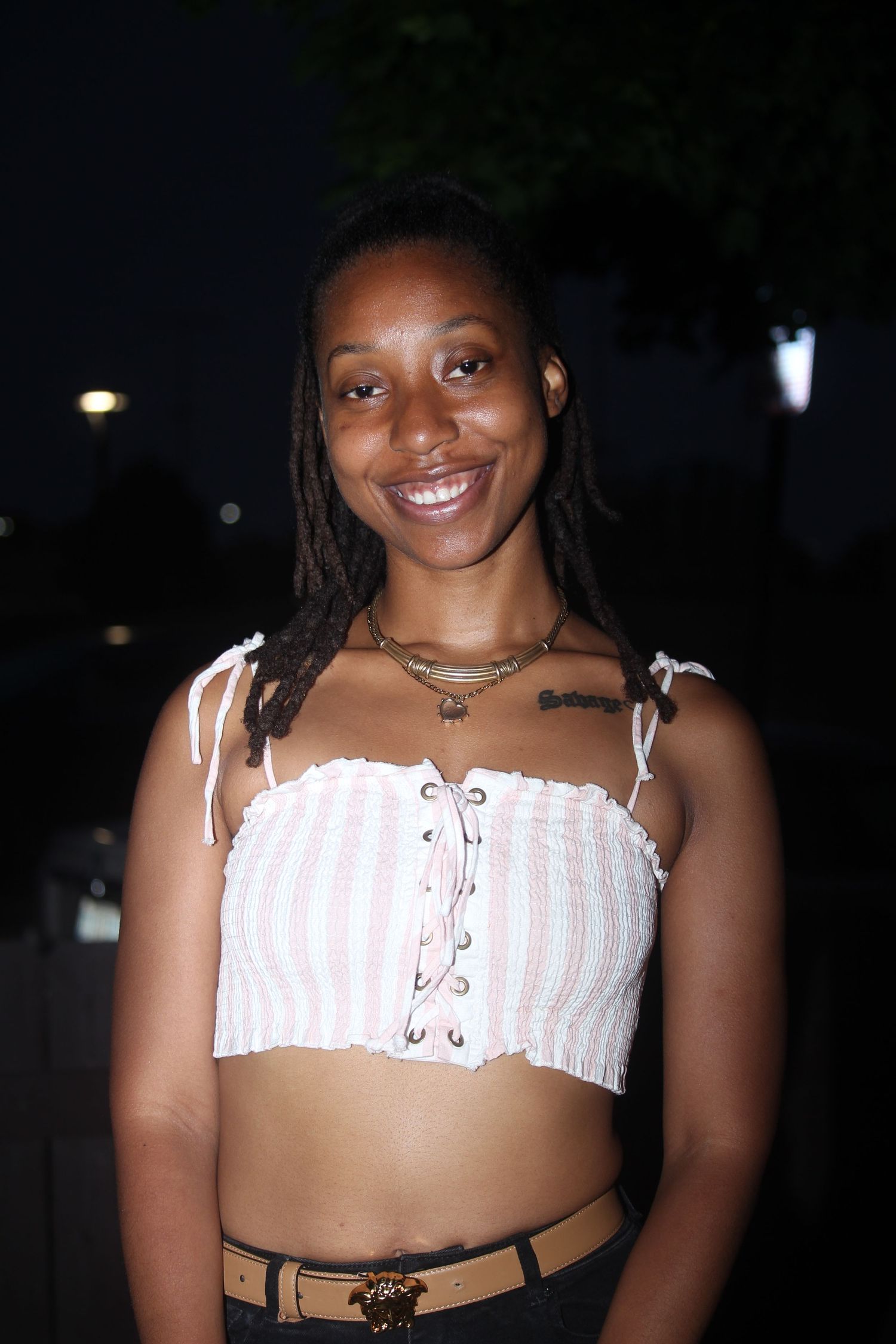 Smiling young woman with locs wearing a striped crop top and belt poses outdoors at night.
