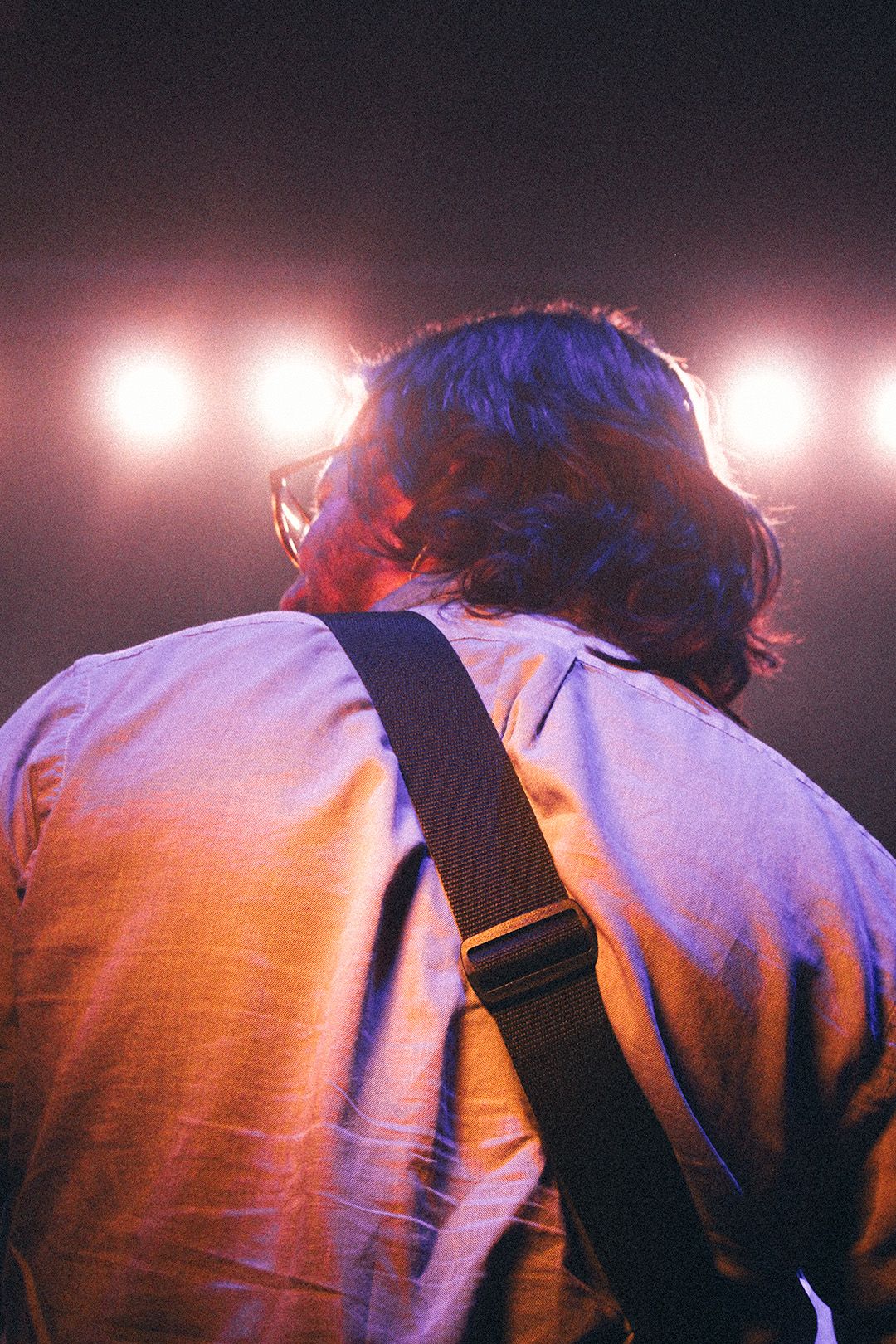 Rear view of long-haired guitarist on stage with bright white spotlights illuminating from behind.