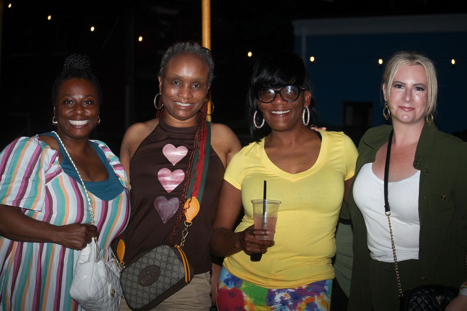 Four women smile together at an outdoor evening event, one holding a drink.