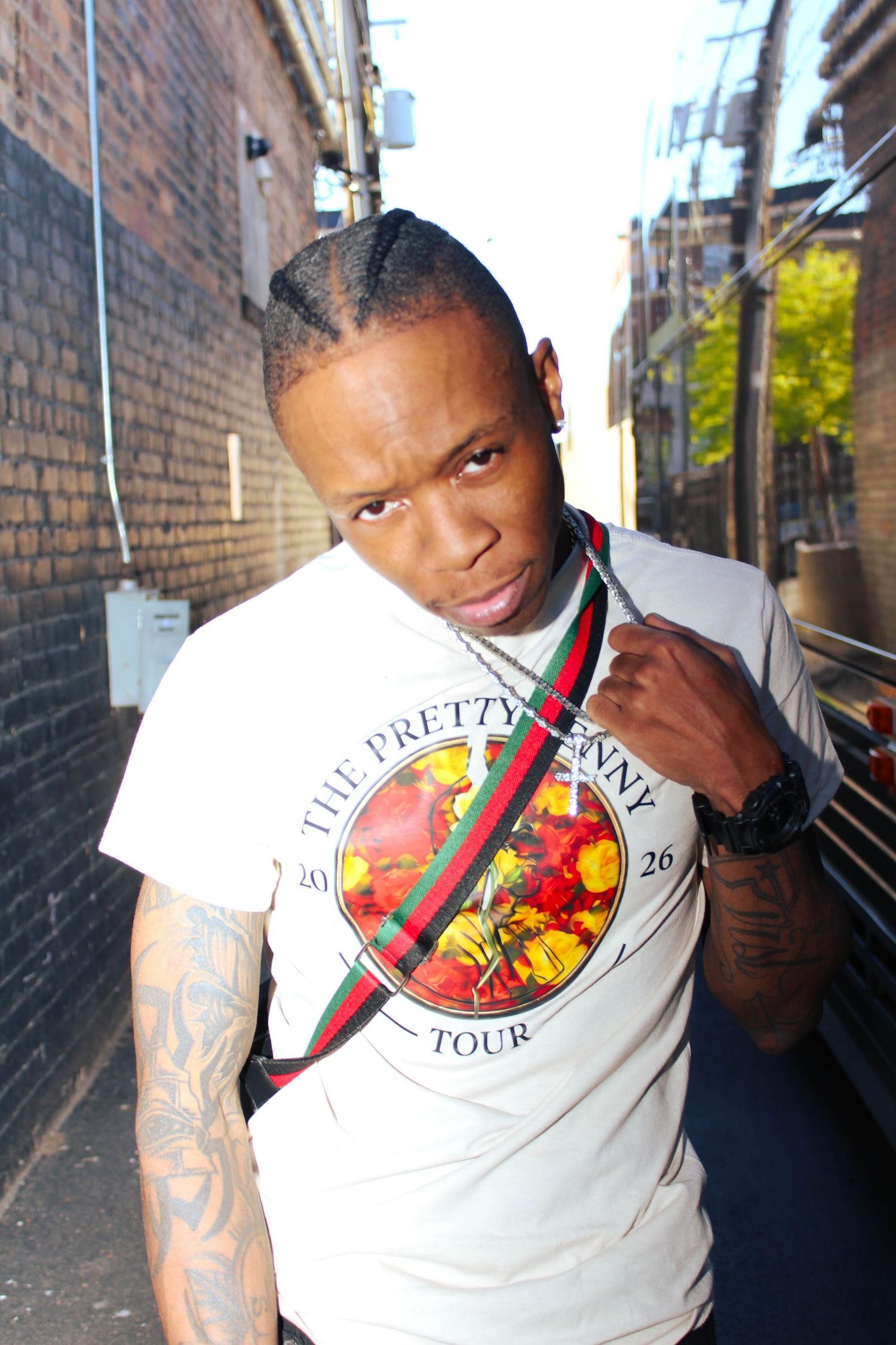 Young man in white Pretty Penny Tour t-shirt with Gucci strap poses in urban brick alley with tattoos visible.