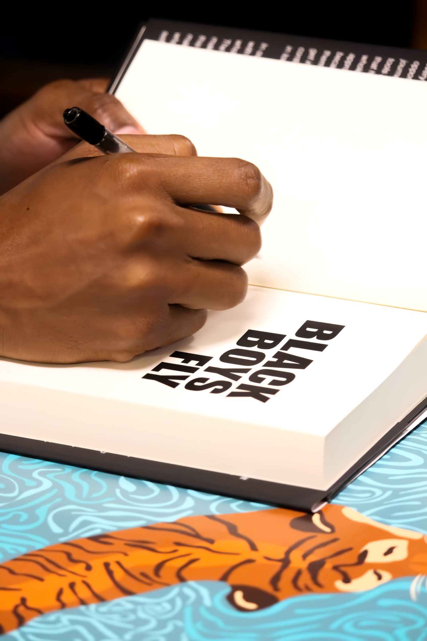 A hand signing the title page of 'Black Boys Fly' book with a pen at a signing event.