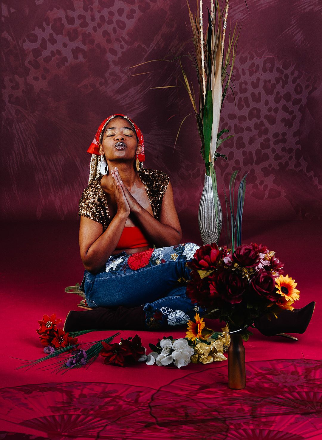 Woman in leopard top and jeans meditates among colorful flowers in red studio setting.