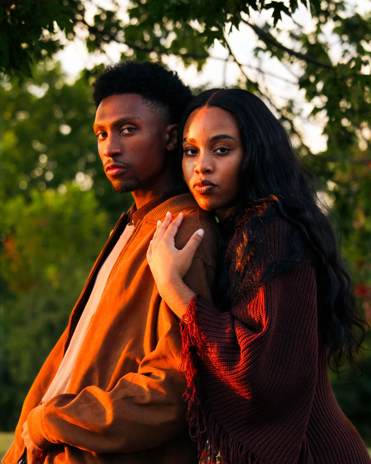 Golden hour portrait of a stylish couple in earth tones posing together under green trees.