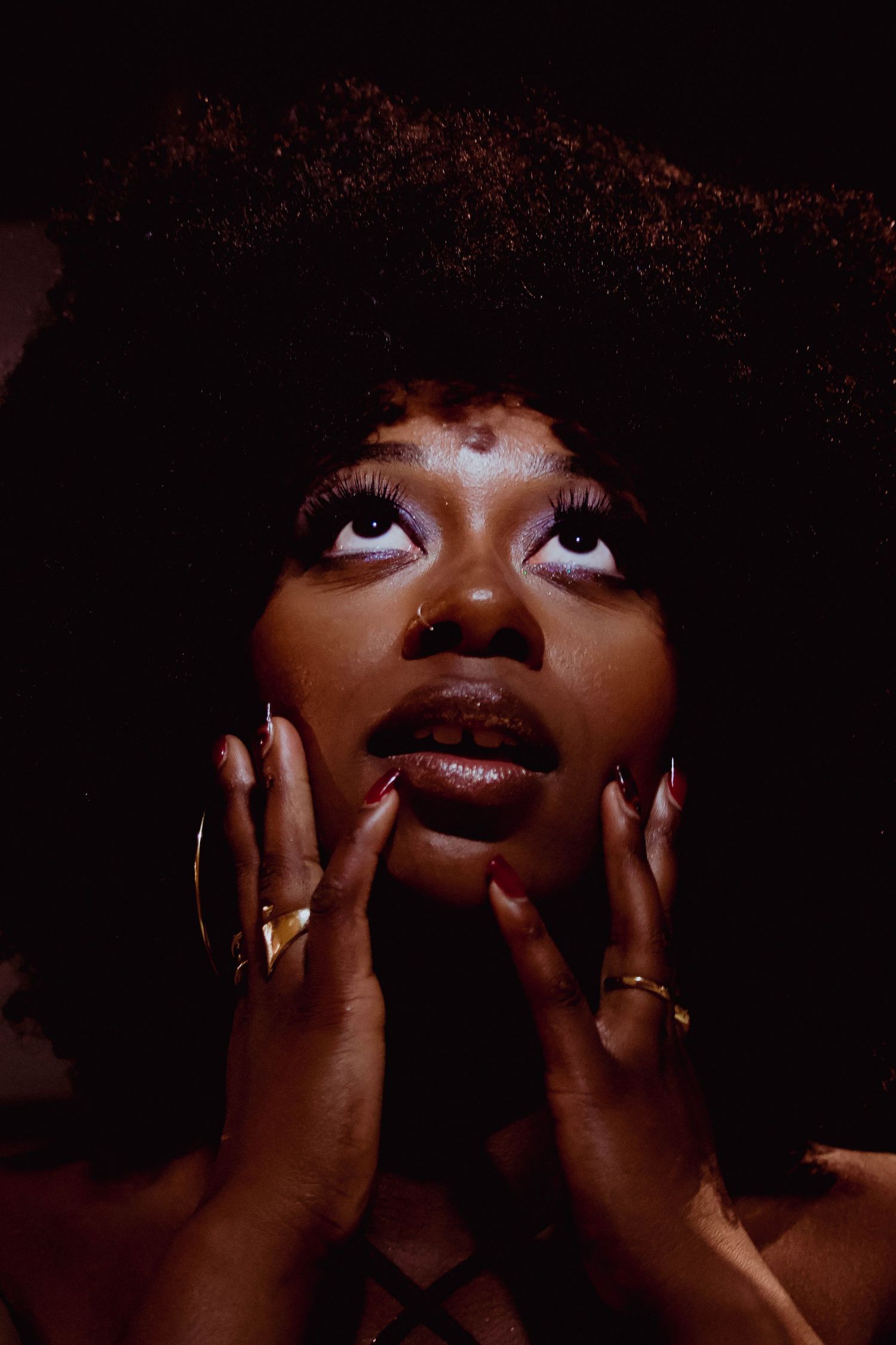 A Black woman with an afro hairstyle poses dramatically with hands framing her face, wearing gold rings and hoop earrings.
