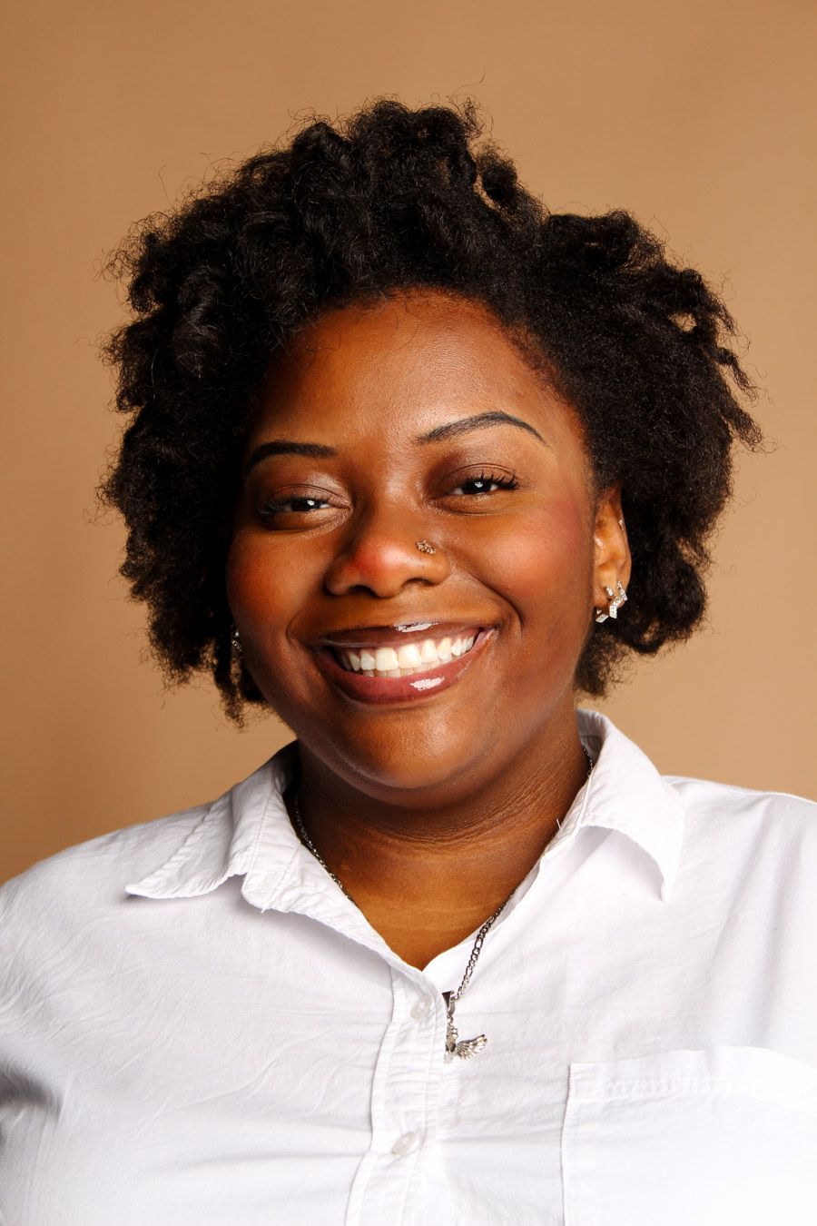 A smiling Black woman with natural hair wears a white button-up shirt against a tan background.