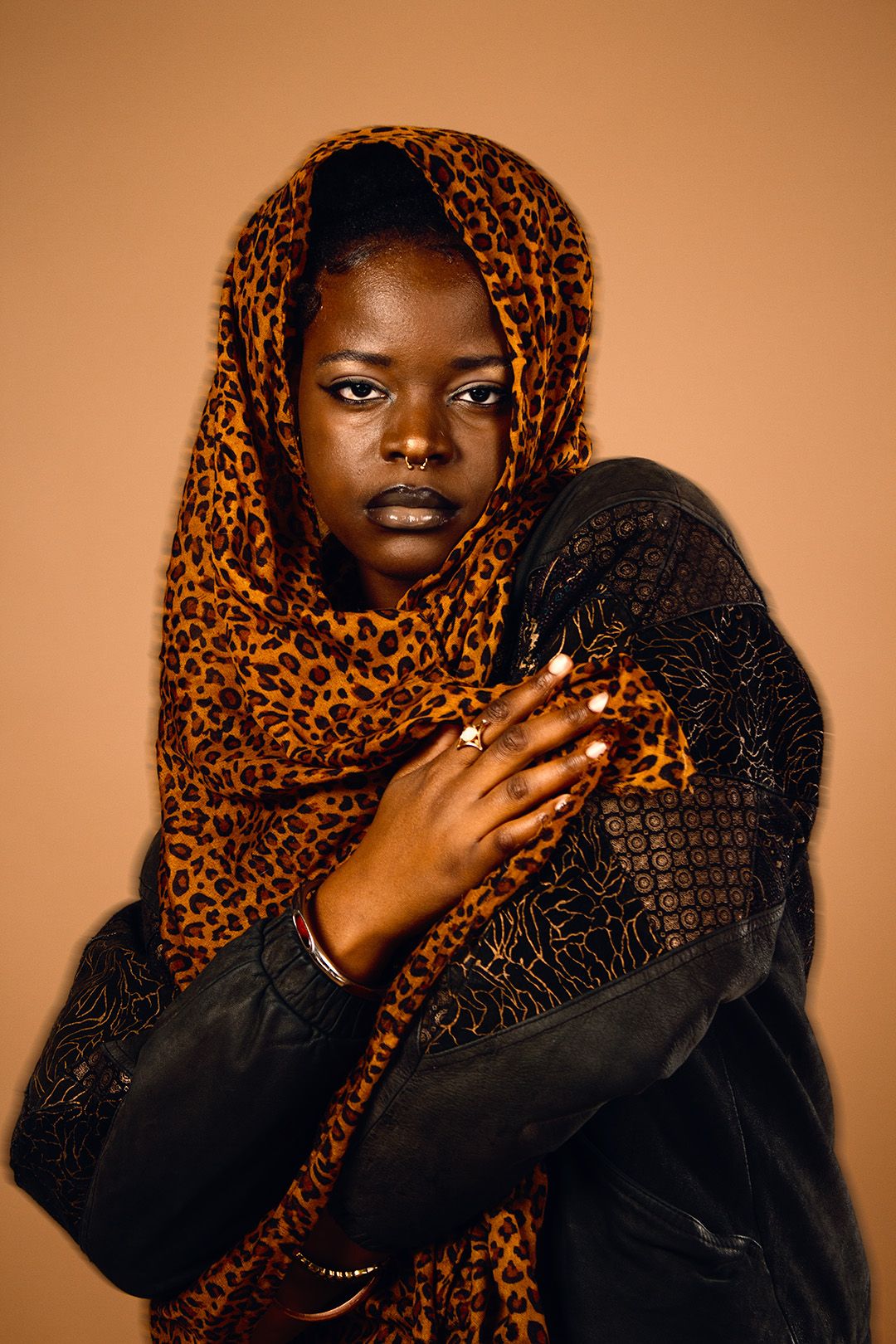 Woman in leopard print headscarf and black beaded jacket posing on a warm beige background.
