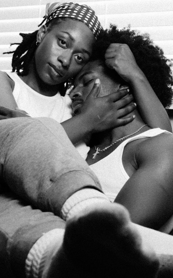Black and white photo of two young Black women sharing an intimate, tender moment while relaxing together indoors.