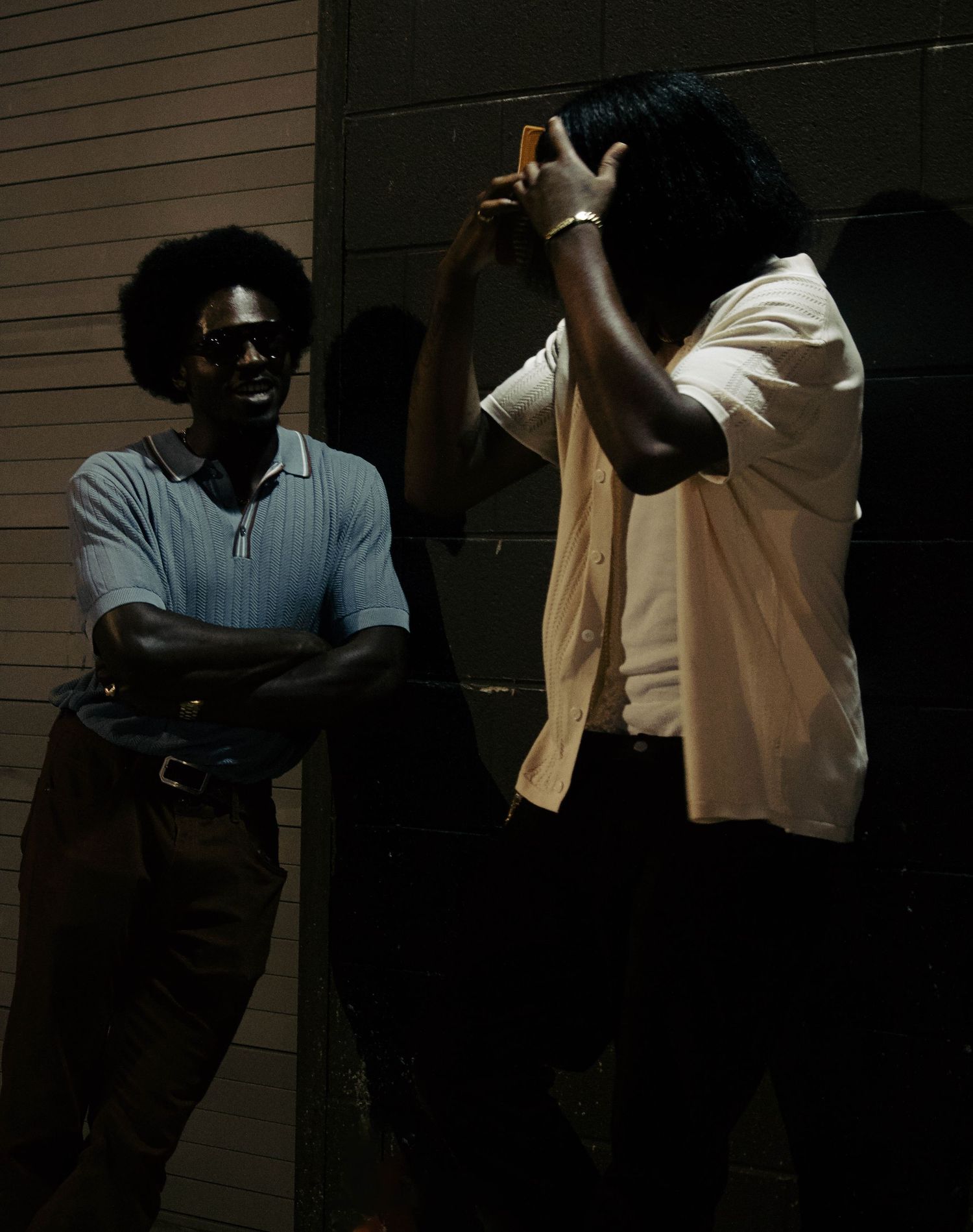 Two men standing against a wall at night, one smiling with arms crossed, the other holding a phone up.