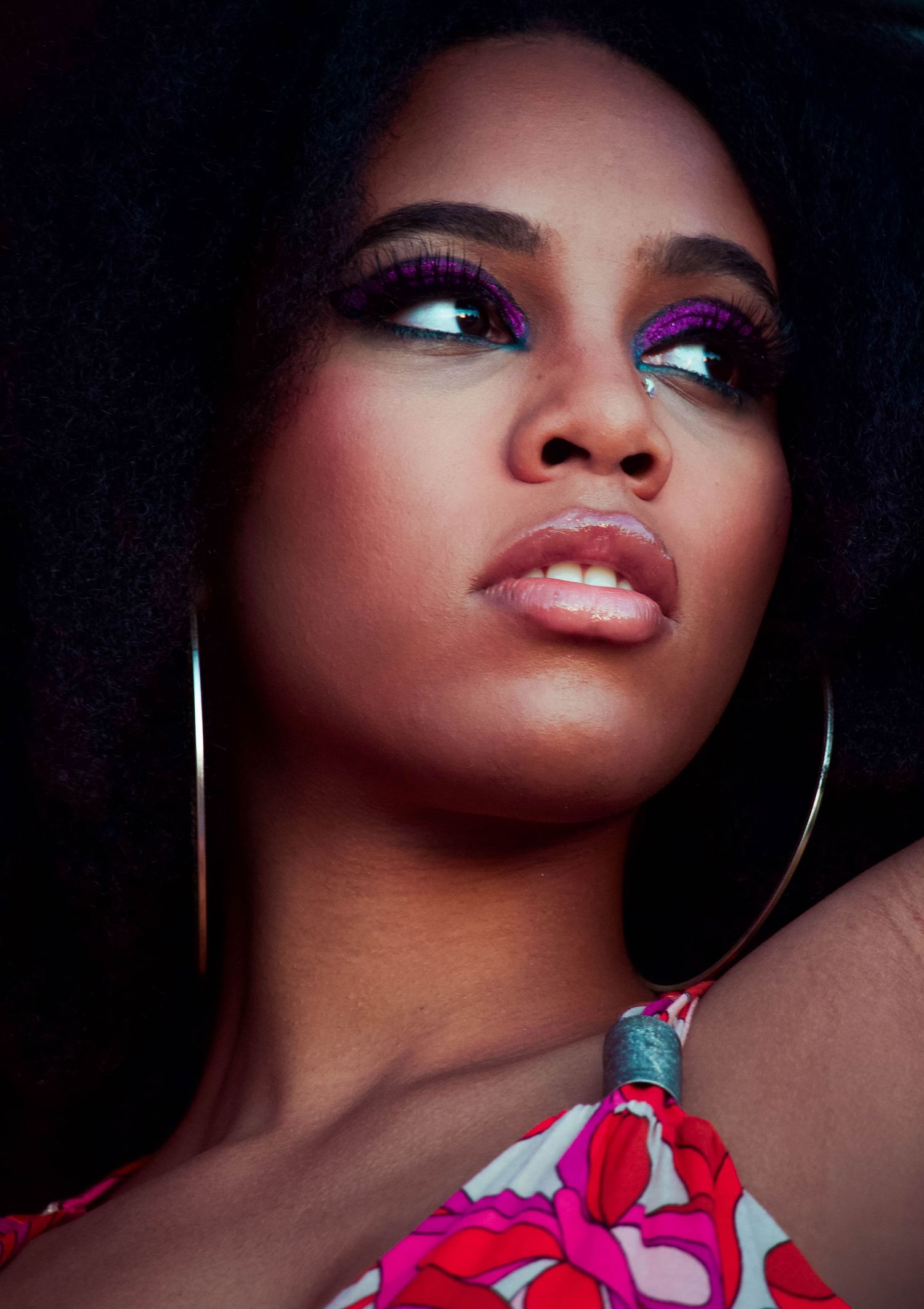 Woman with bold purple eyeshadow, hoop earrings, and floral top poses elegantly against dark background.