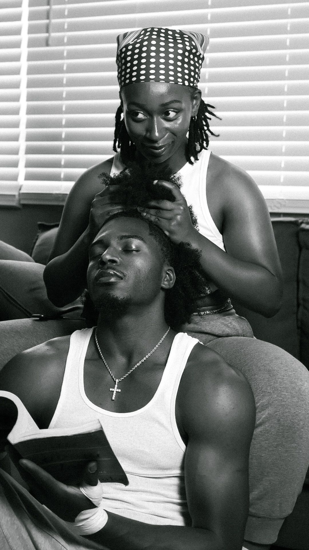 A black and white photo of a couple relaxing on a sofa, woman braiding mans hair.
