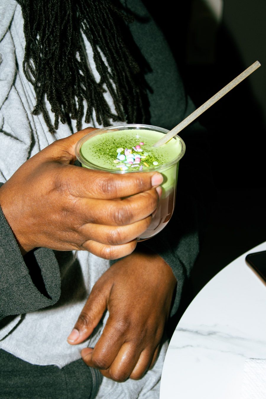 Person with dreadlocks holding a green matcha latte topped with flower petals.