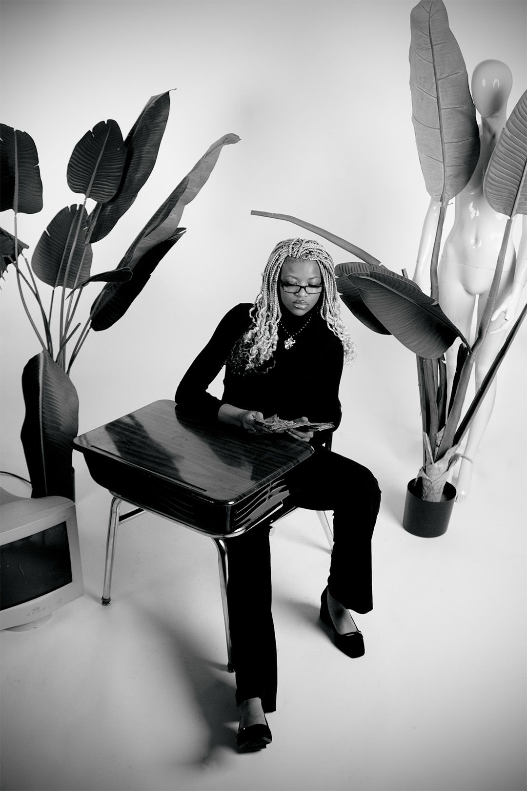 A Black woman with blonde locs wearing glasses sits on a desk surrounded by tropical plants in black and white.