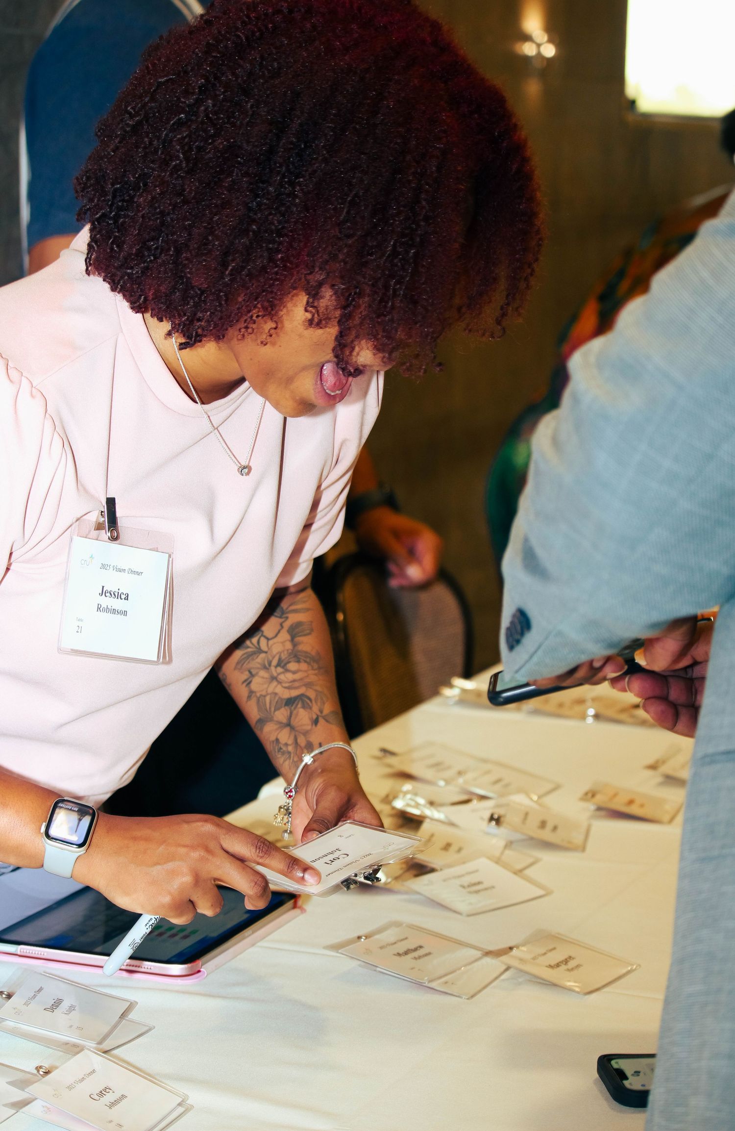 Woman with red curly hair and tattoos checking name badges at an event registration table.