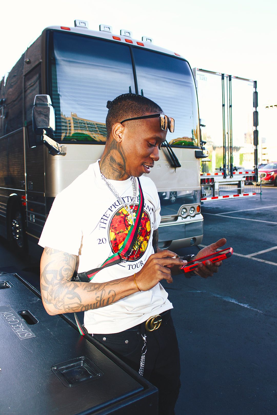 Tattooed man in white tour t-shirt using phone near large tour bus in parking lot.