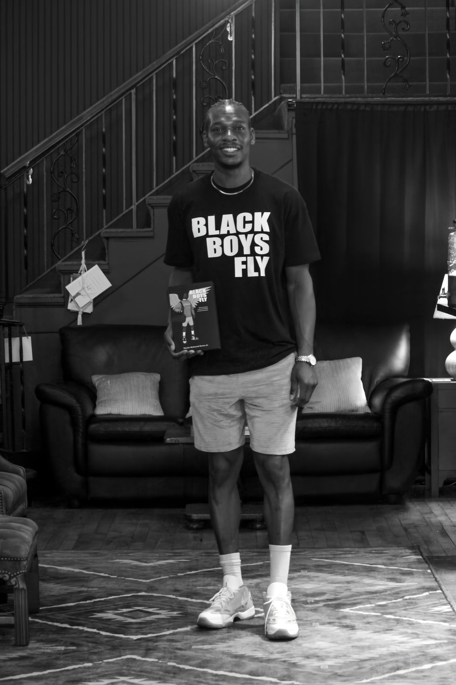 Man in Black Boys Fly t-shirt and shorts holding a book, standing in front of a leather couch and staircase.