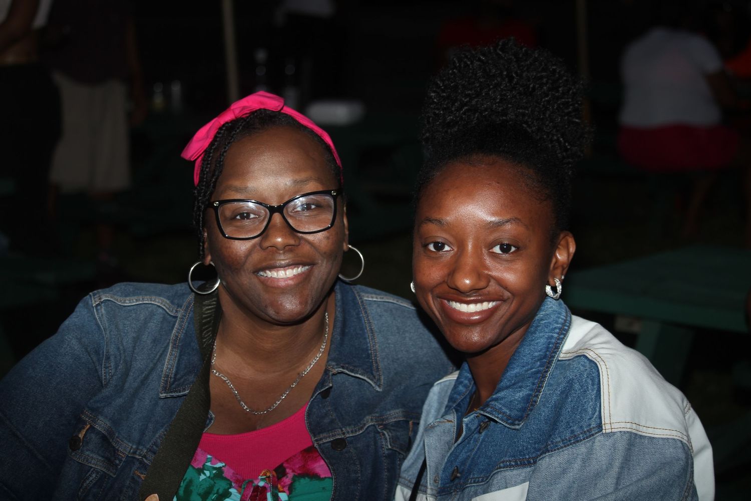 Two smiling women in denim jackets pose together at an outdoor nighttime gathering.