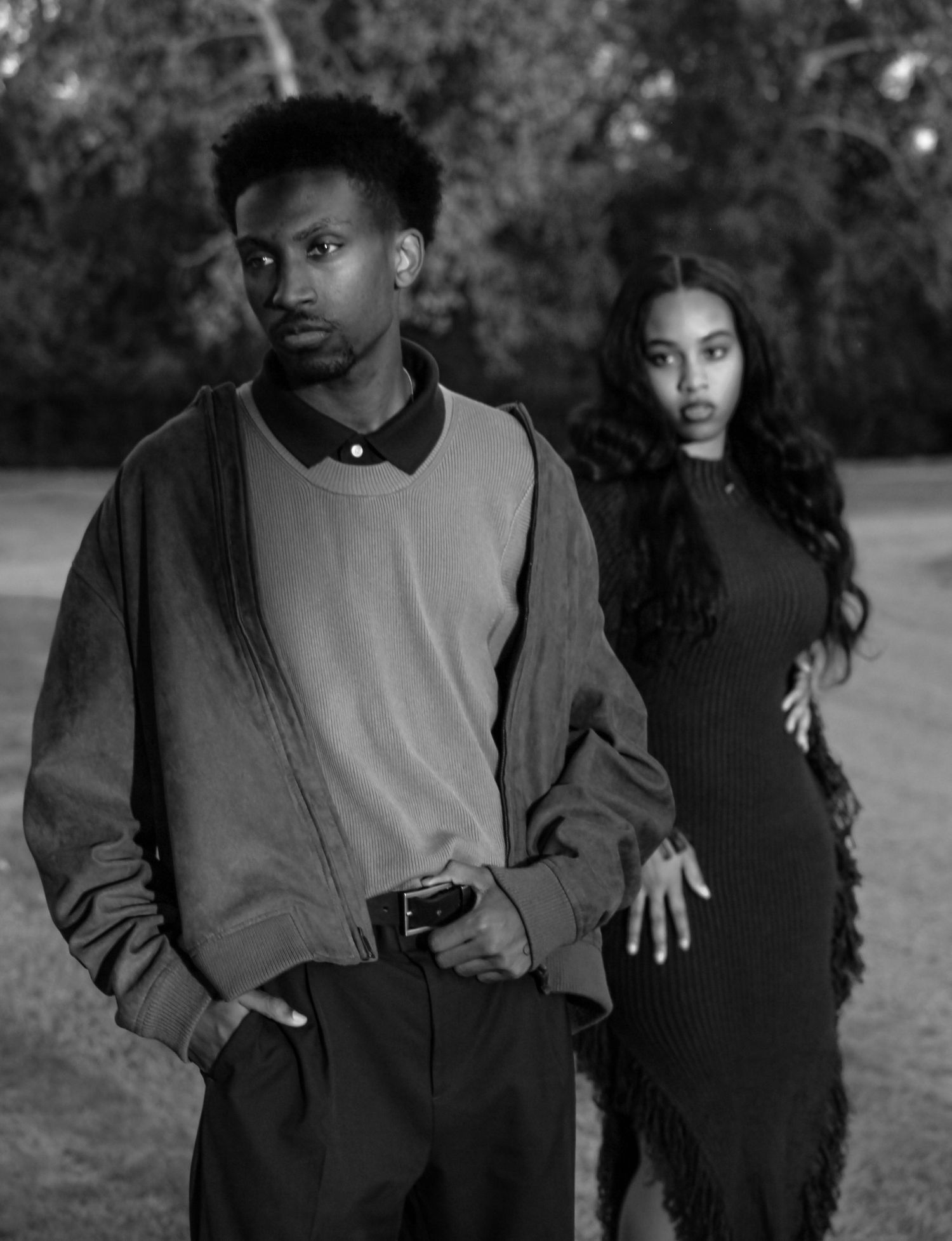 Black and white outdoor photo of a young couple standing apart in casual and elegant attire.