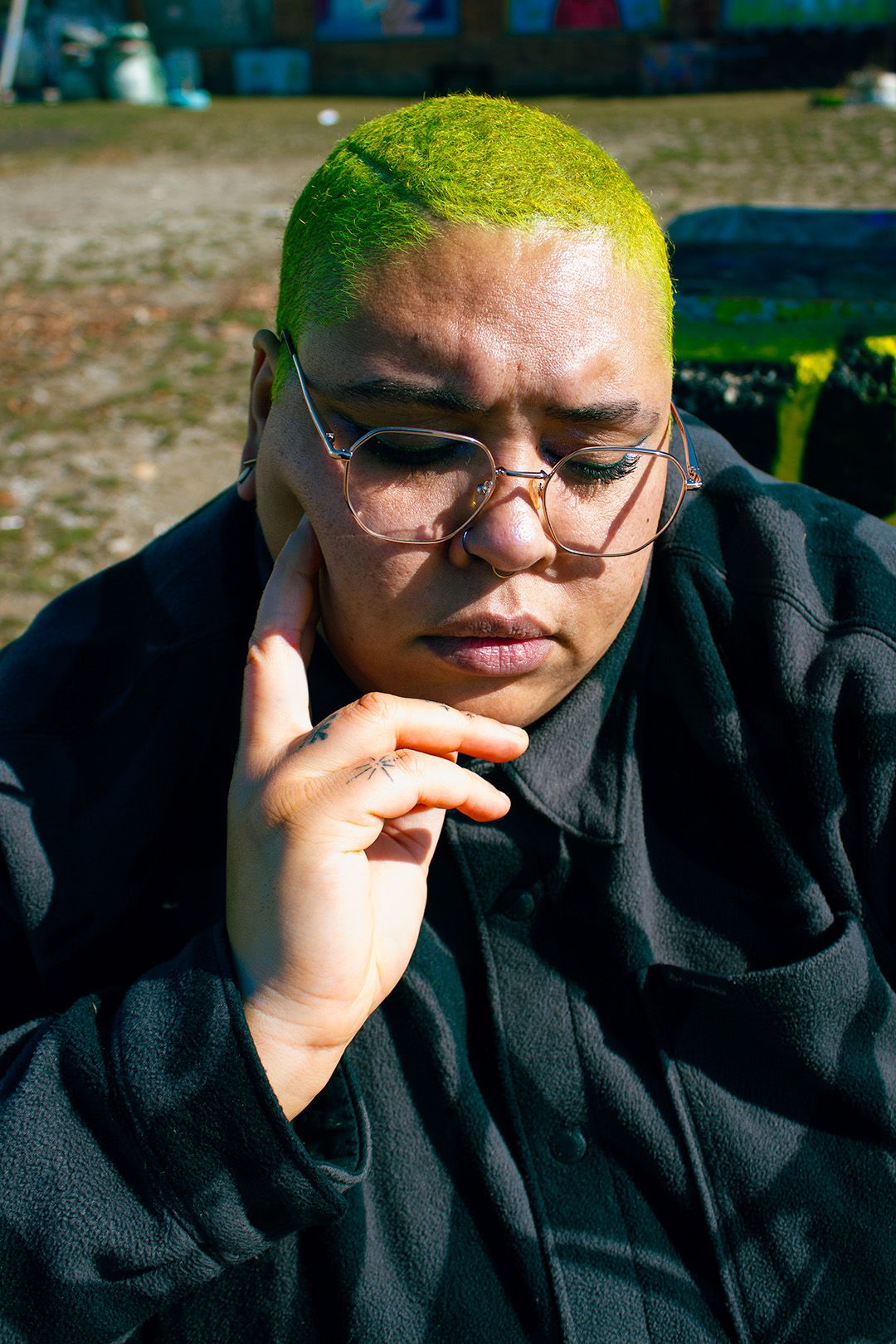 Person with bright green buzz cut and glasses resting chin on hand outdoors.