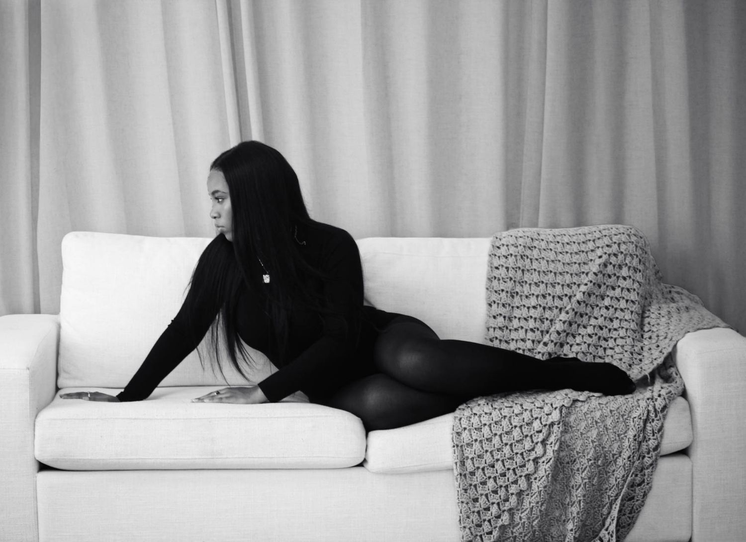 Black and white photo of a woman in a black outfit reclining on a white sofa with a knit blanket.