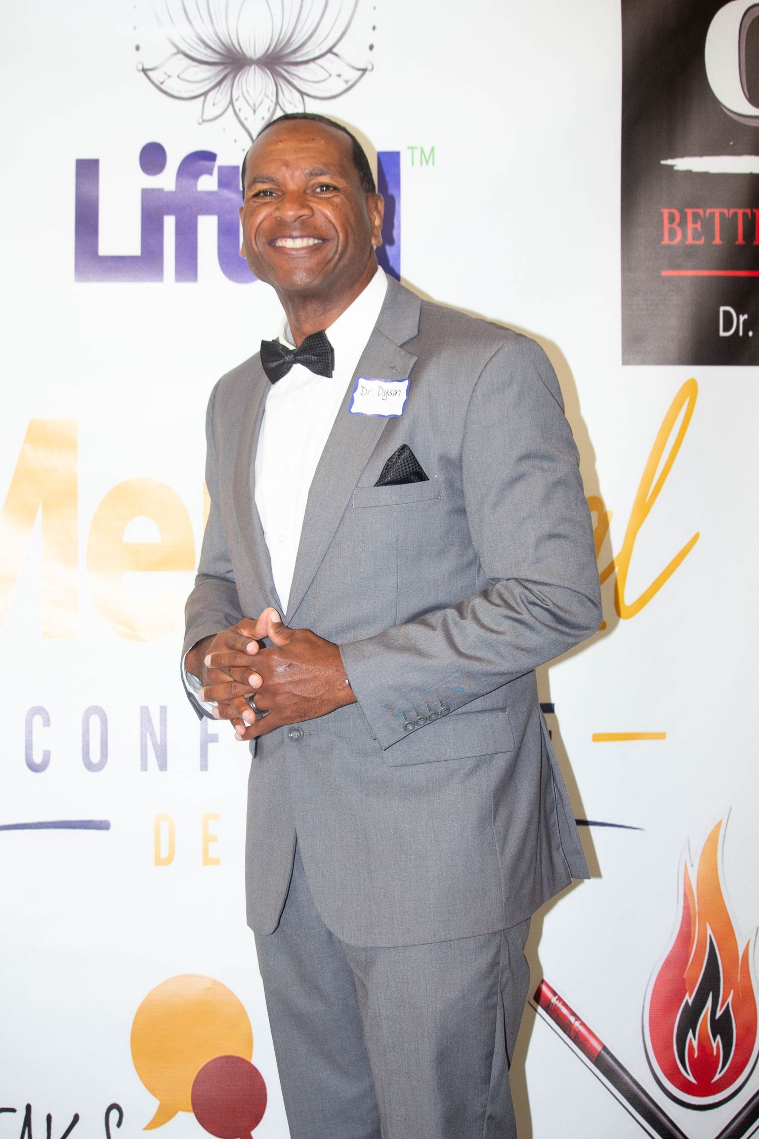 Man in gray tuxedo with bow tie smiling at Mental Health Conference Detroit event on white step-and-repeat backdrop.