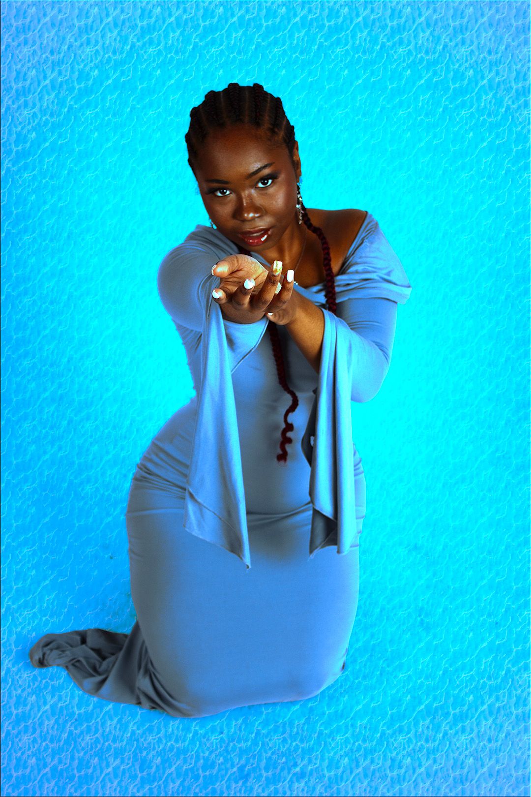 Black woman in blue dress kneels and points toward camera with manicured nails against blue background.