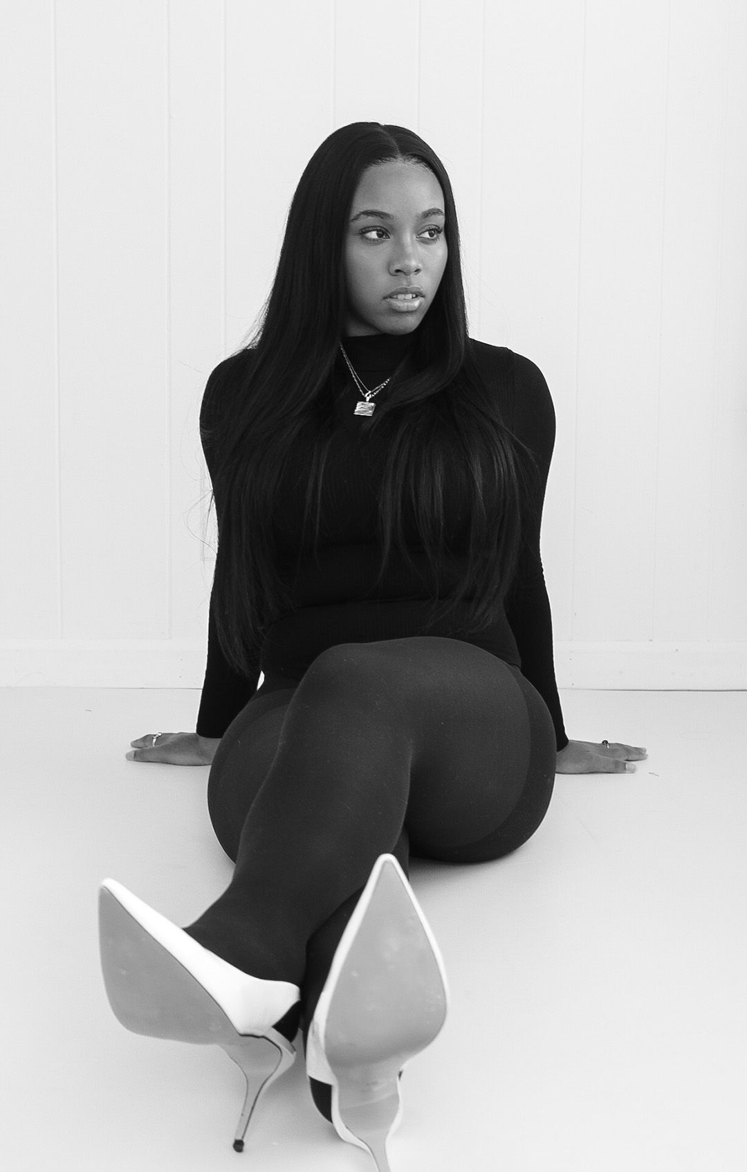 Black and white photo of a young woman in black outfit and white heels sitting on floor posing.