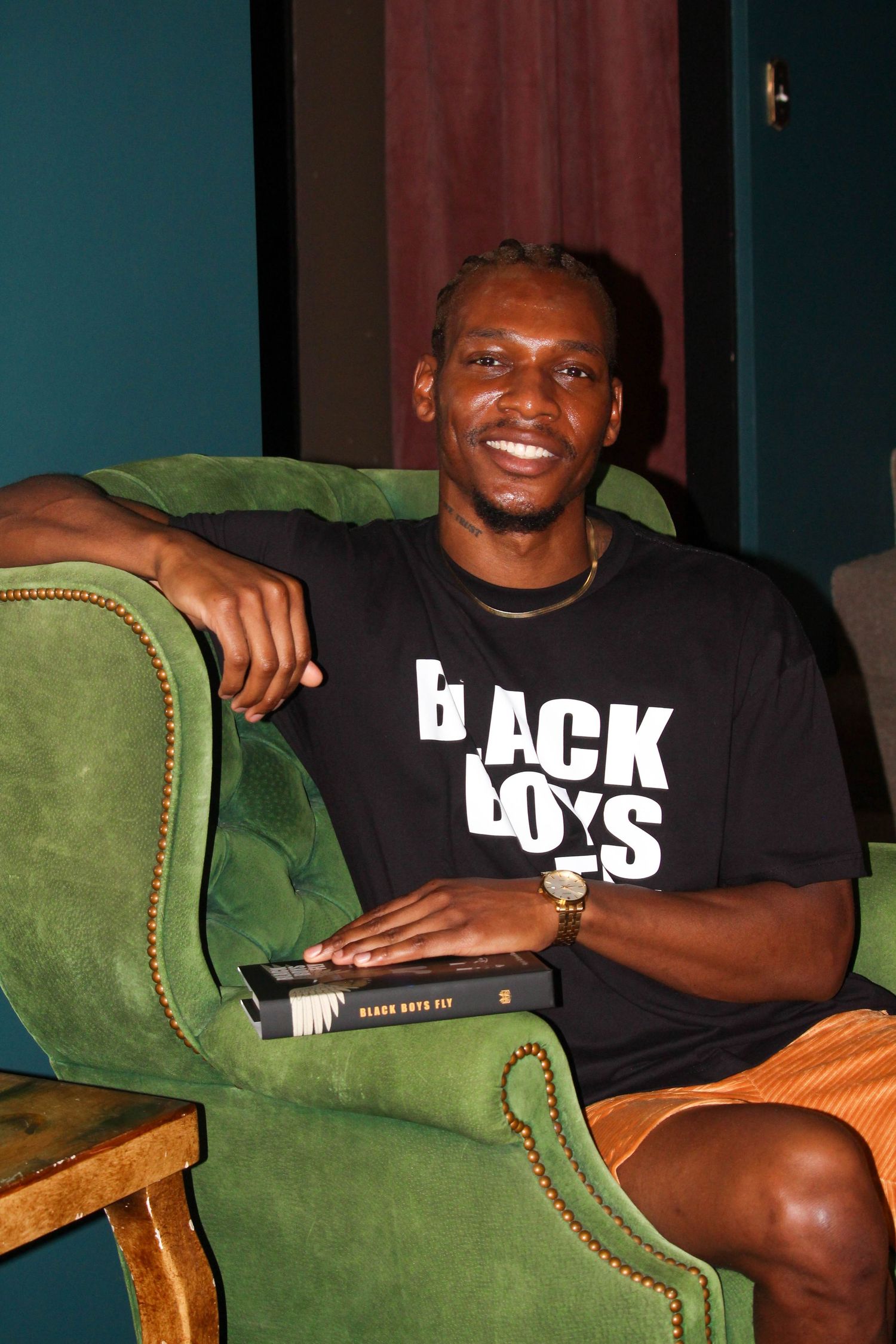 Man in Black Boys Fly t-shirt sits smiling in green armchair holding a book.