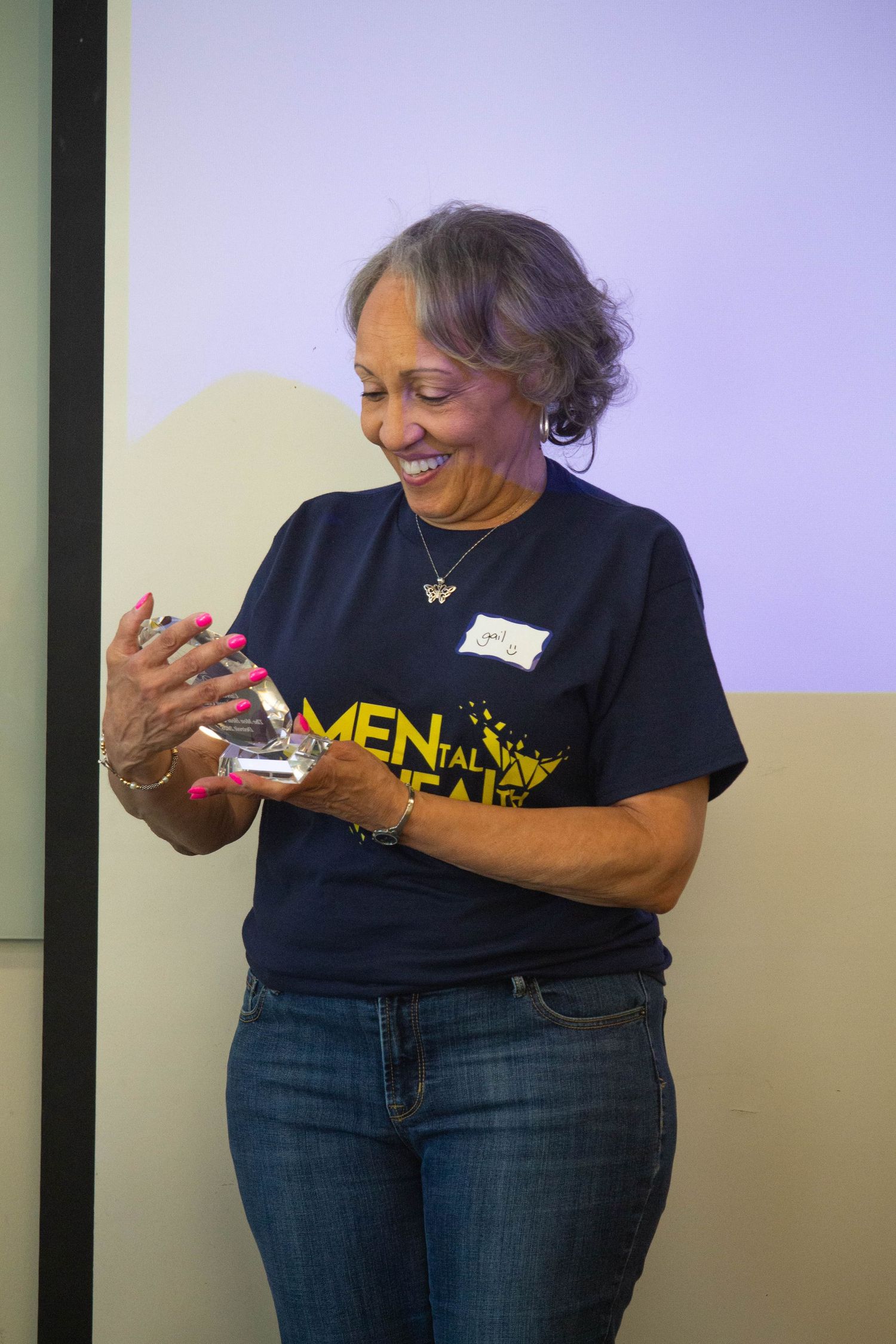 Smiling woman in Mental Health t-shirt holding crystal award trophy at community recognition event.