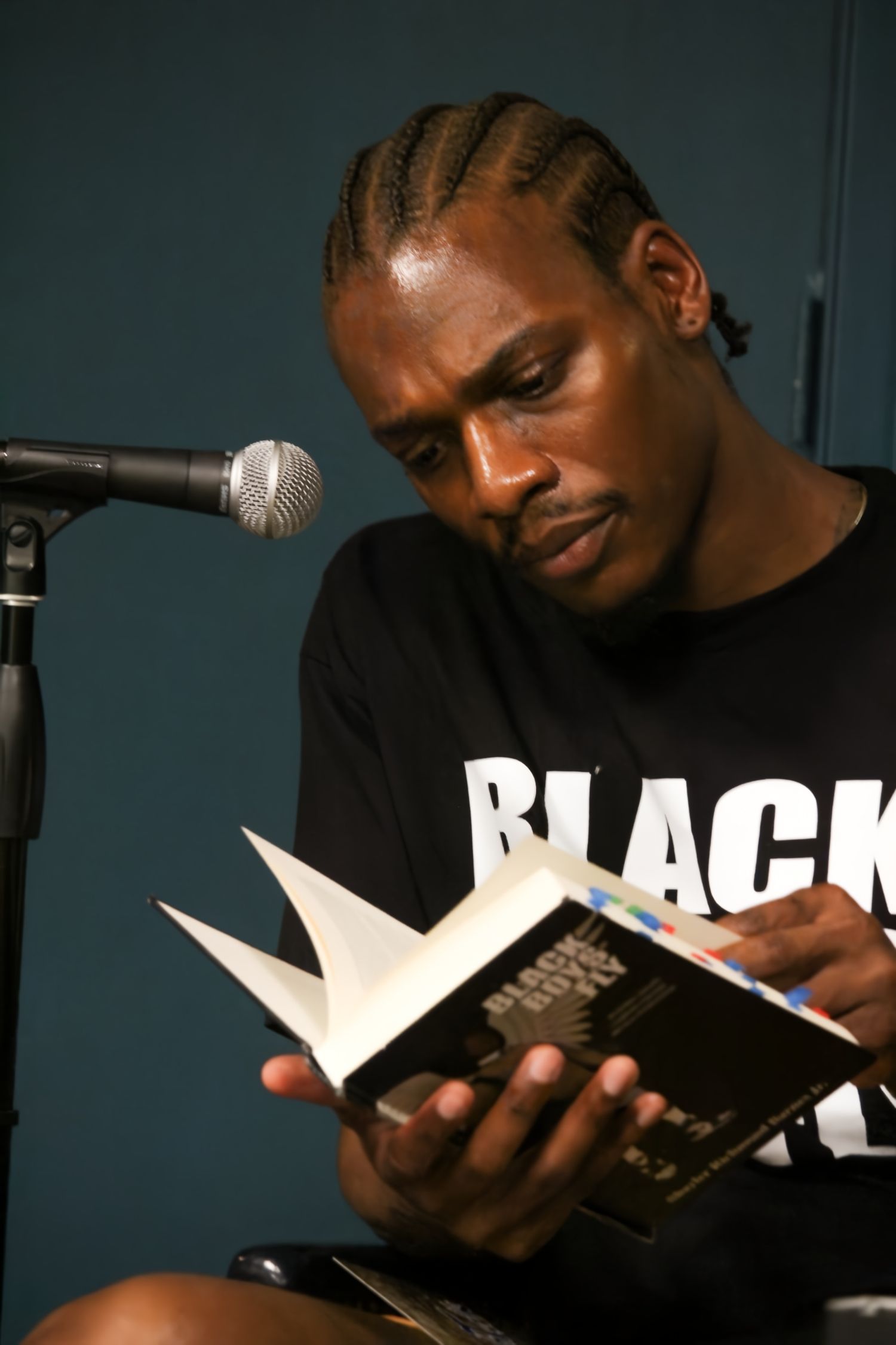 Man with cornrow braids reading a book at a microphone wearing a black t-shirt.