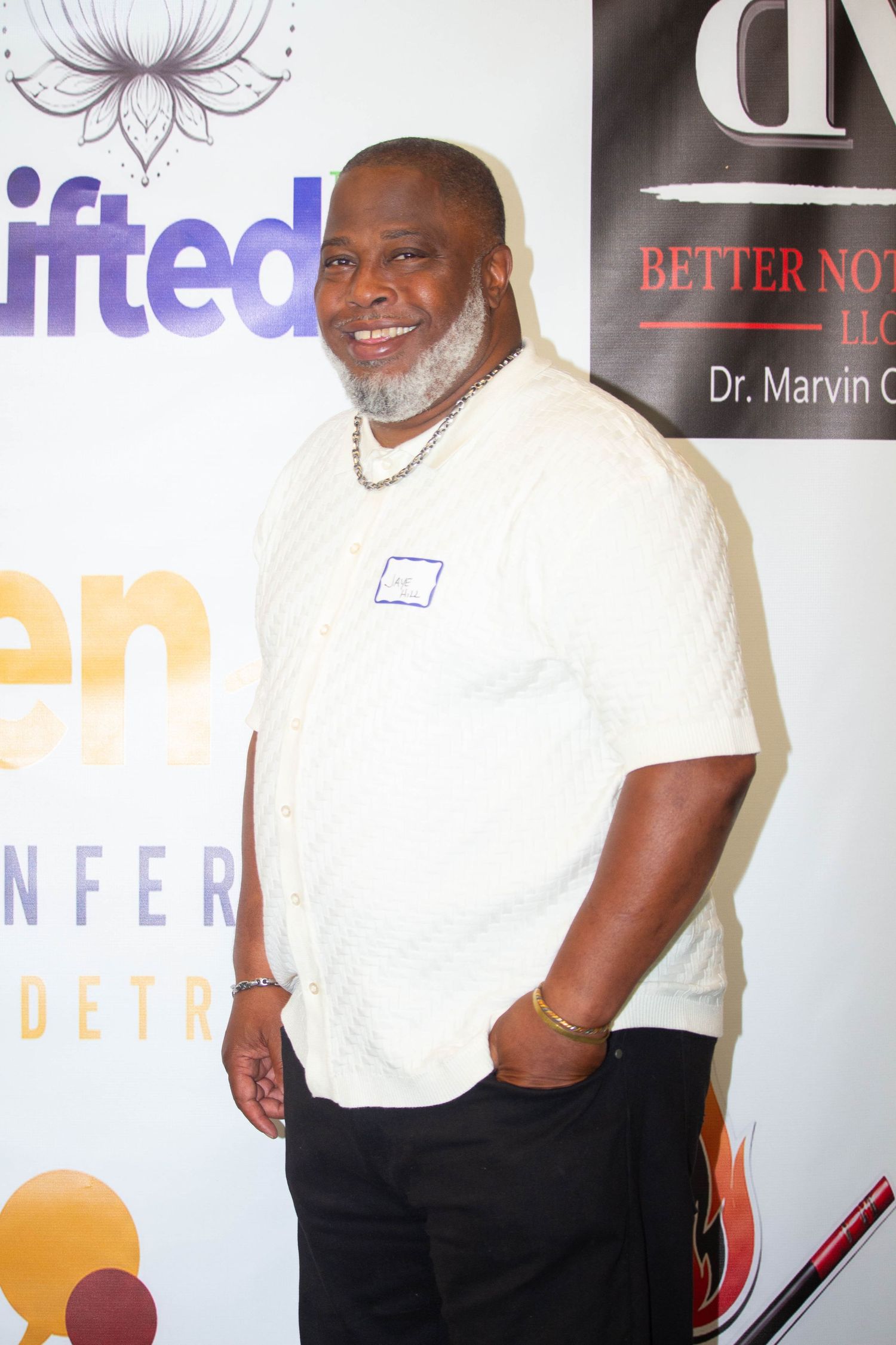 A smiling man in a white knit shirt and black pants poses at an event backdrop featuring sponsor logos.